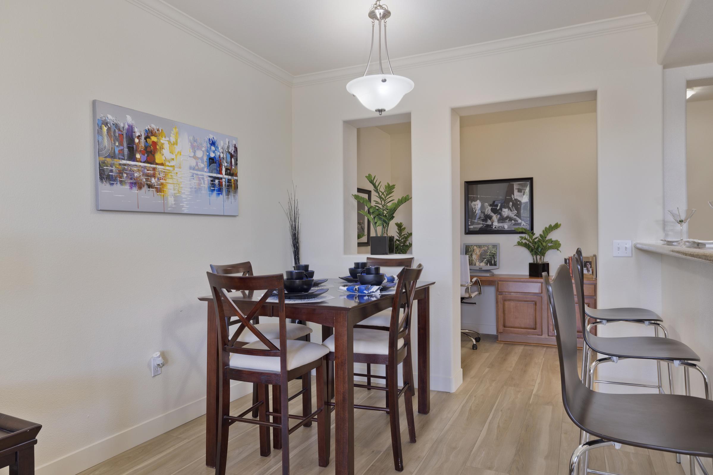 A modern dining area featuring a wooden dining table with four chairs, decorated with black plates. In the background, a small nook with an office setup and a potted plant. A colorful abstract painting hangs on the wall, alongside soft lighting from a pendant lamp. The floor is light-colored wood, contributing to a bright, inviting atmosphere.