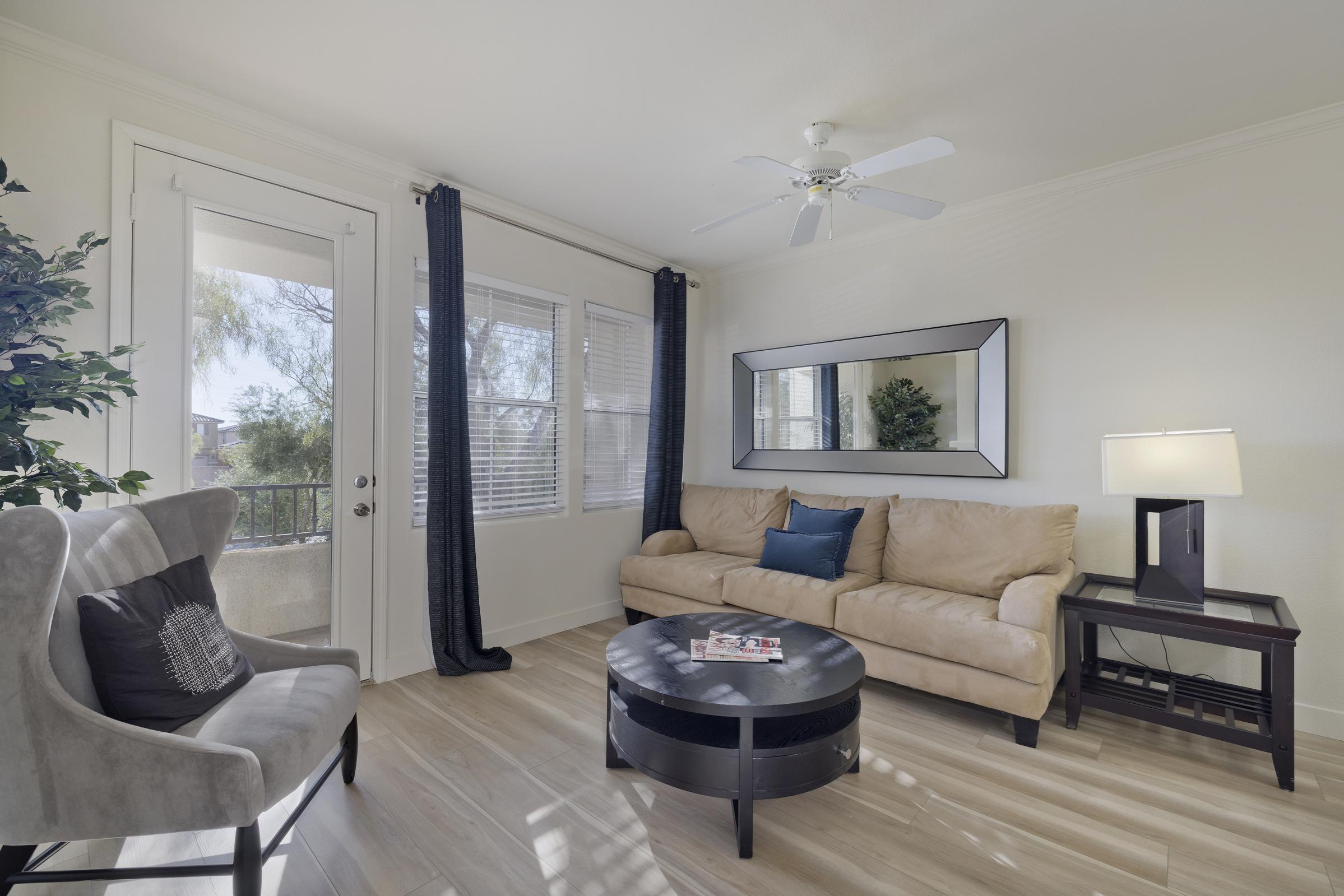 A cozy living room featuring a light-colored sofa with a blue decorative pillow, a round black coffee table with magazines, and a modern armchair. There is a large mirror on the wall, a lamp on a side table, and a door leading to a balcony with a view of greenery. Natural light fills the space through the windows.