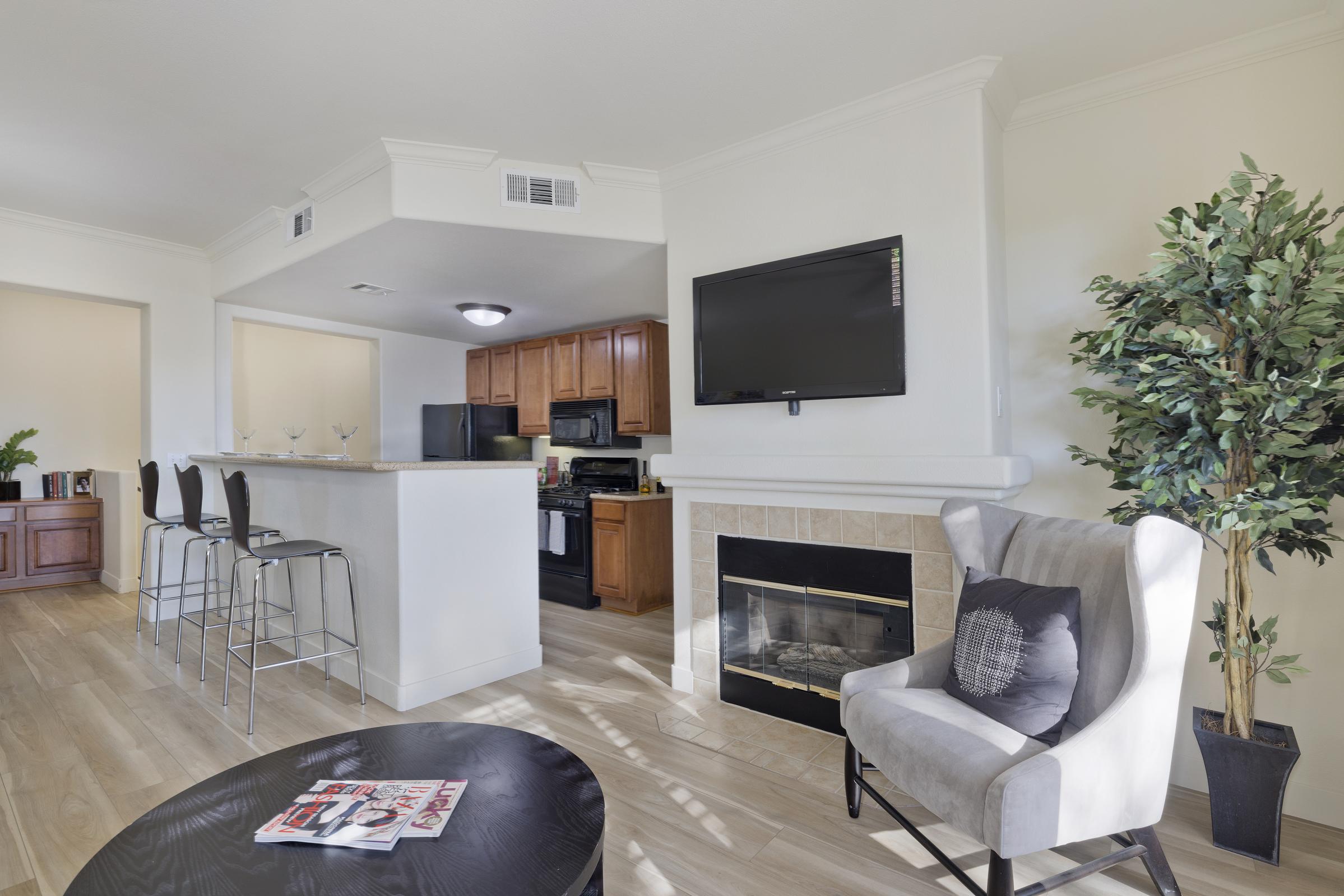 A modern living space featuring a kitchen with wooden cabinets and a black stove, a bar area with tall stools, a cozy fireplace, and a stylish chair. The room is bright with natural light, and decorative plants add a touch of greenery. A coffee table holds a magazine.