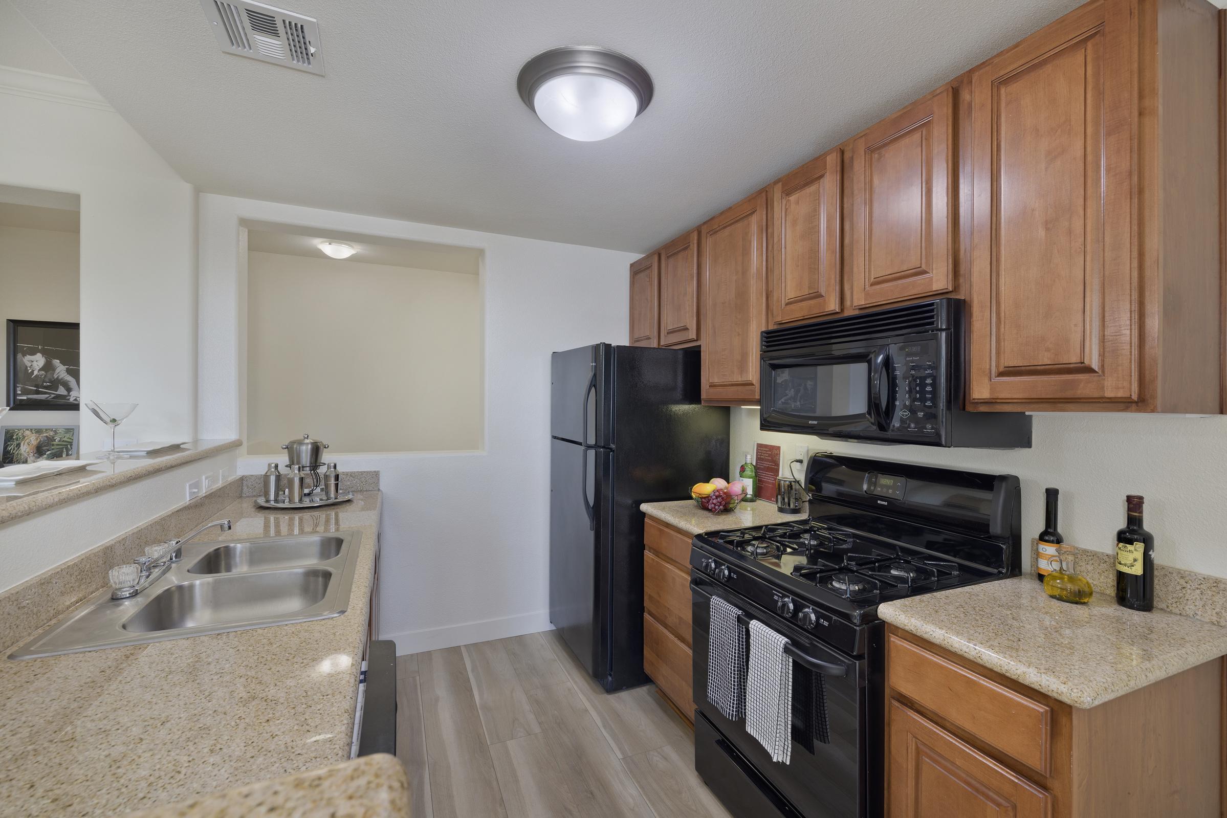 Modern kitchen with wooden cabinets, granite countertops, a black refrigerator, and a black gas stove. There are dish soap and a dish rack by the sink, along with a fruit bowl and wine bottles on the counter. The space features a bright ceiling light and has an open layout.