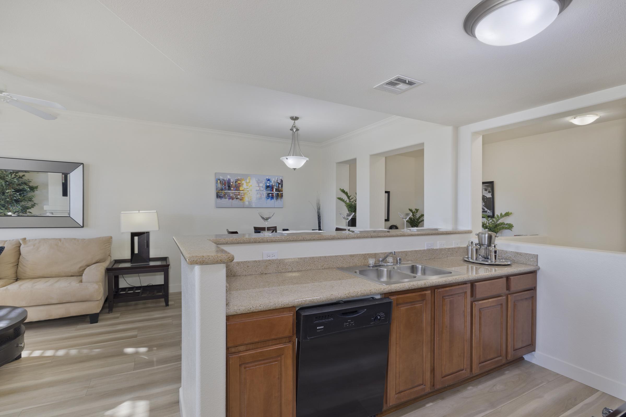 A modern kitchen featuring wood cabinets, a double sink, and a black dishwasher. In the background, a light-filled living area with a beige couch, a lamp, and decorative artwork on the wall. The space includes potted plants, creating a warm and inviting atmosphere.