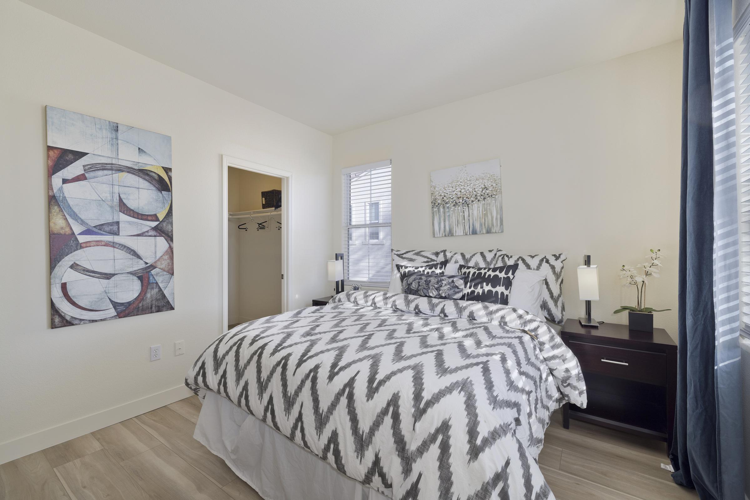A cozy bedroom featuring a gray and white chevron-patterned bedspread on a queen-sized bed, nightstands with lamps on either side, and a colorful abstract painting on the wall. A closet door is visible in the background, with natural light coming through the windows.