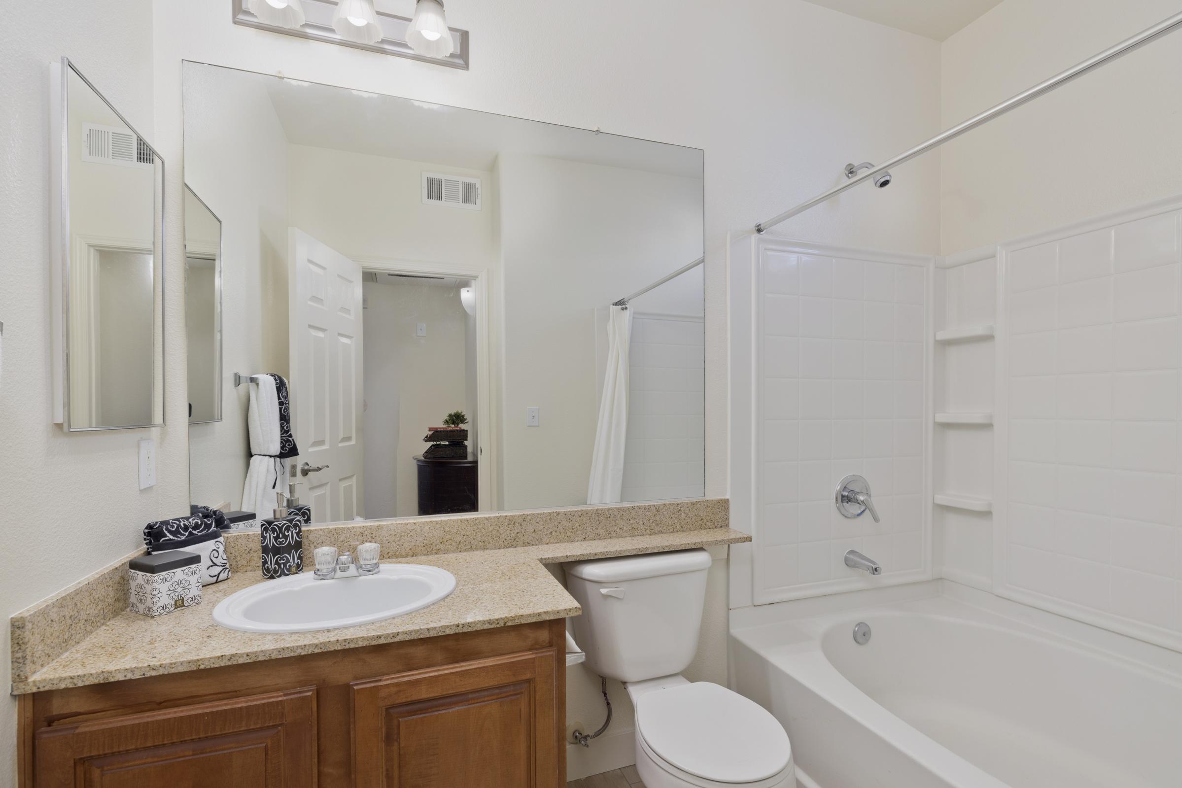 A modern bathroom featuring a light-colored wall, a large mirror above a granite countertop with a sink, a white toilet, and a bathtub with a shower curtain. Shelving is visible in the bathtub area, and a door leads to another room in the background. Decorative towels are neatly arranged.