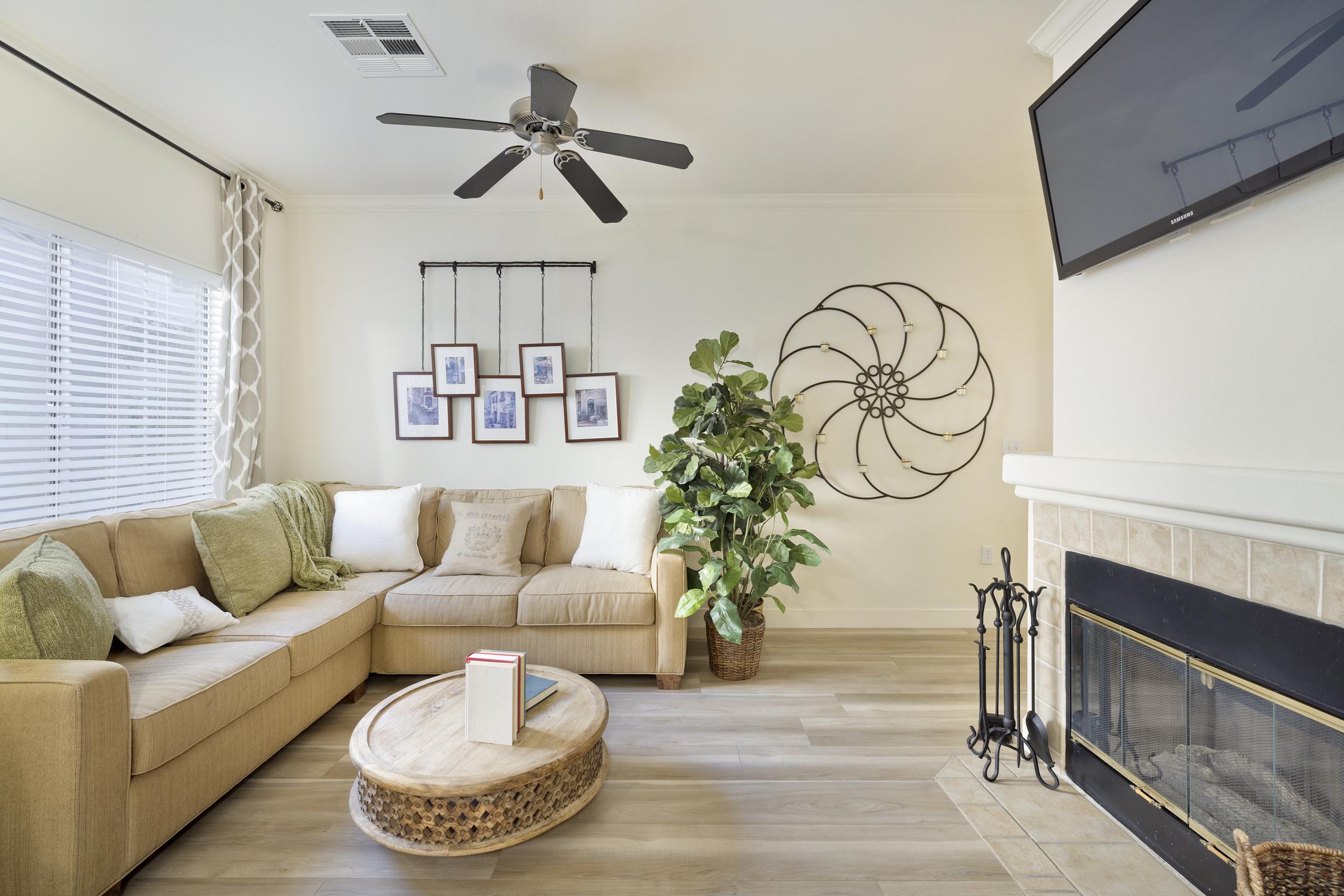 A cozy living room featuring a light brown sectional sofa with green and white throw pillows, a round coffee table, and a decorative plant. The wall has framed photos, a spiral wall decoration, and a television mounted above a fireplace adorned with a decorative screen. Natural light filters through a window with blinds.