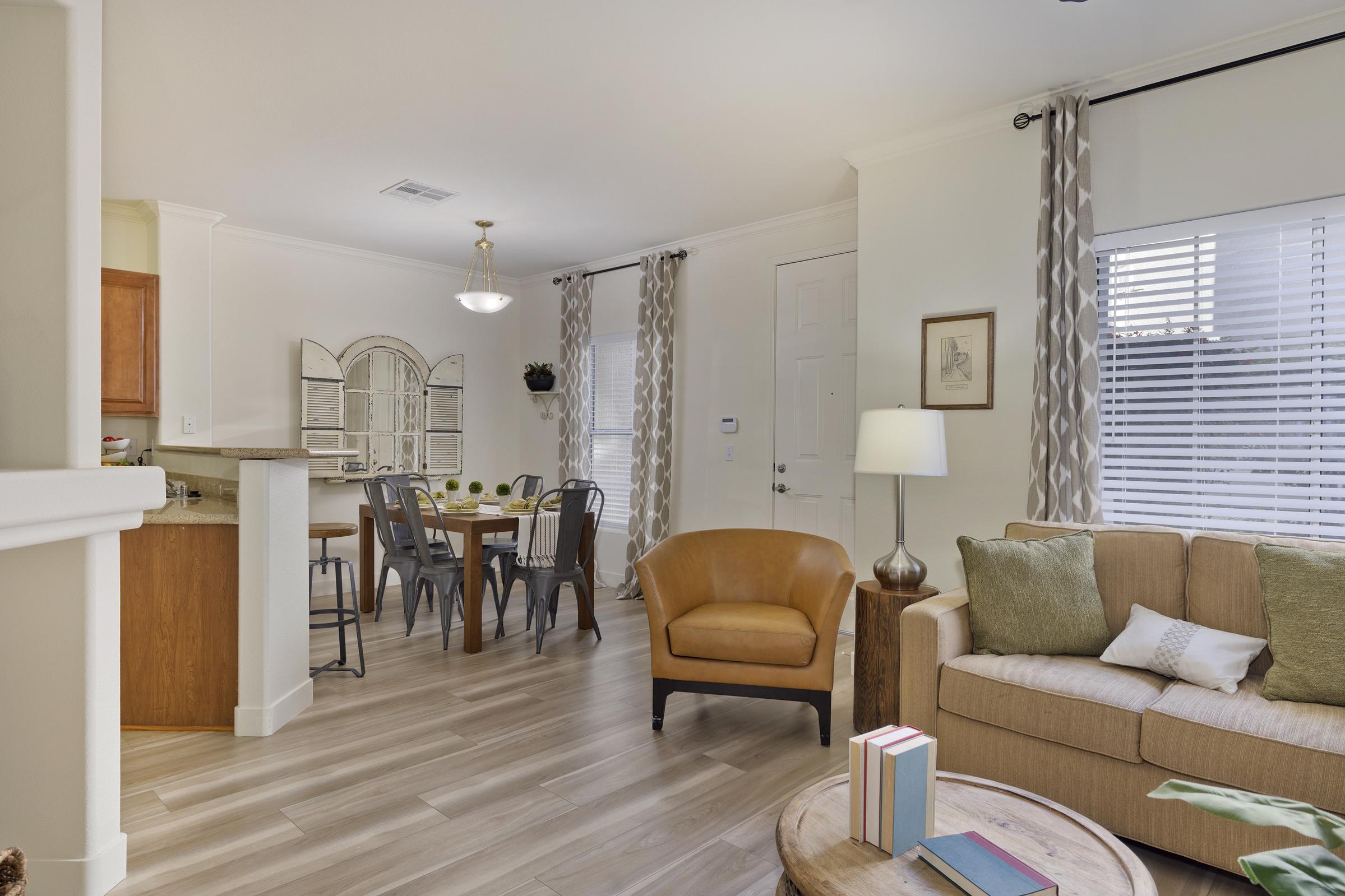 A cozy living room and dining area featuring a brown armchair, a beige sofa adorned with cushions, a coffee table with books, and a dining table with six chairs. Large windows with curtains allow natural light to fill the space, creating a warm and inviting atmosphere.