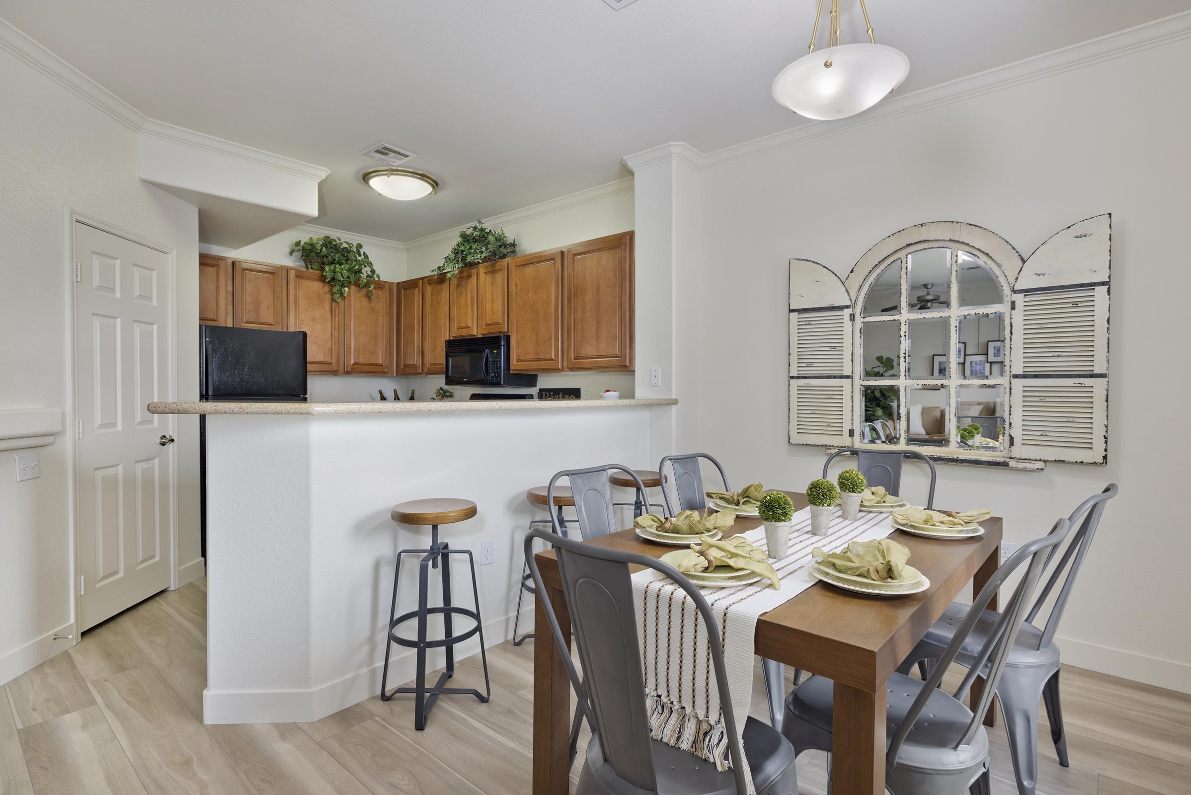 A modern kitchen with light wood cabinetry and a breakfast bar containing tall stools. A dining table is set for four, featuring decorative plates and green accents. Natural light enters through a window styled with shutters, complemented by indoor plants on the countertop and shelves.
