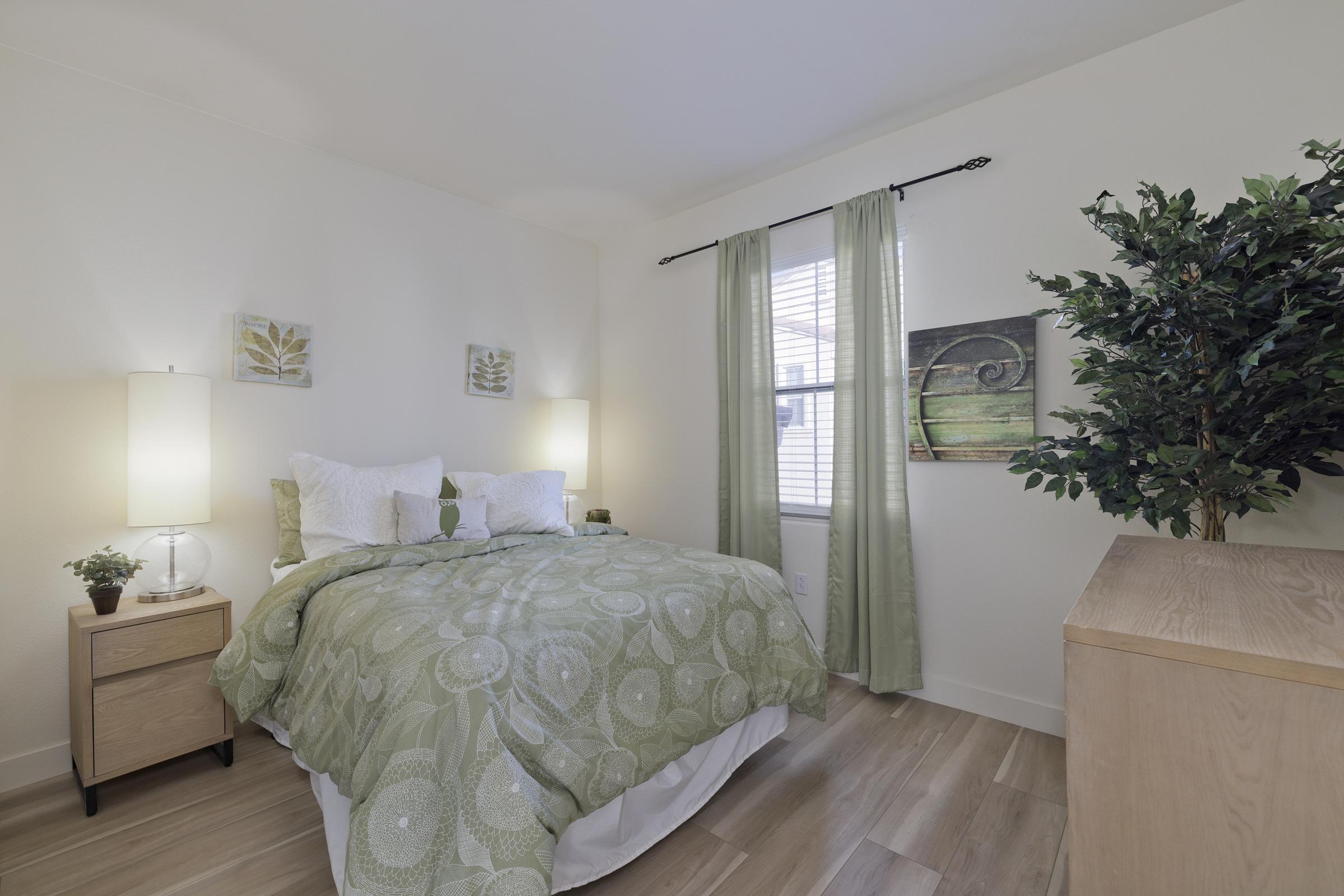Cozy bedroom featuring a green and white bedding set, two bedside lamps, and a wooden nightstand. A potted plant is in the corner, and decorative wall art is displayed above the bed. Natural light enters through a window, complemented by light green curtains. The flooring is wooden, creating a warm atmosphere.