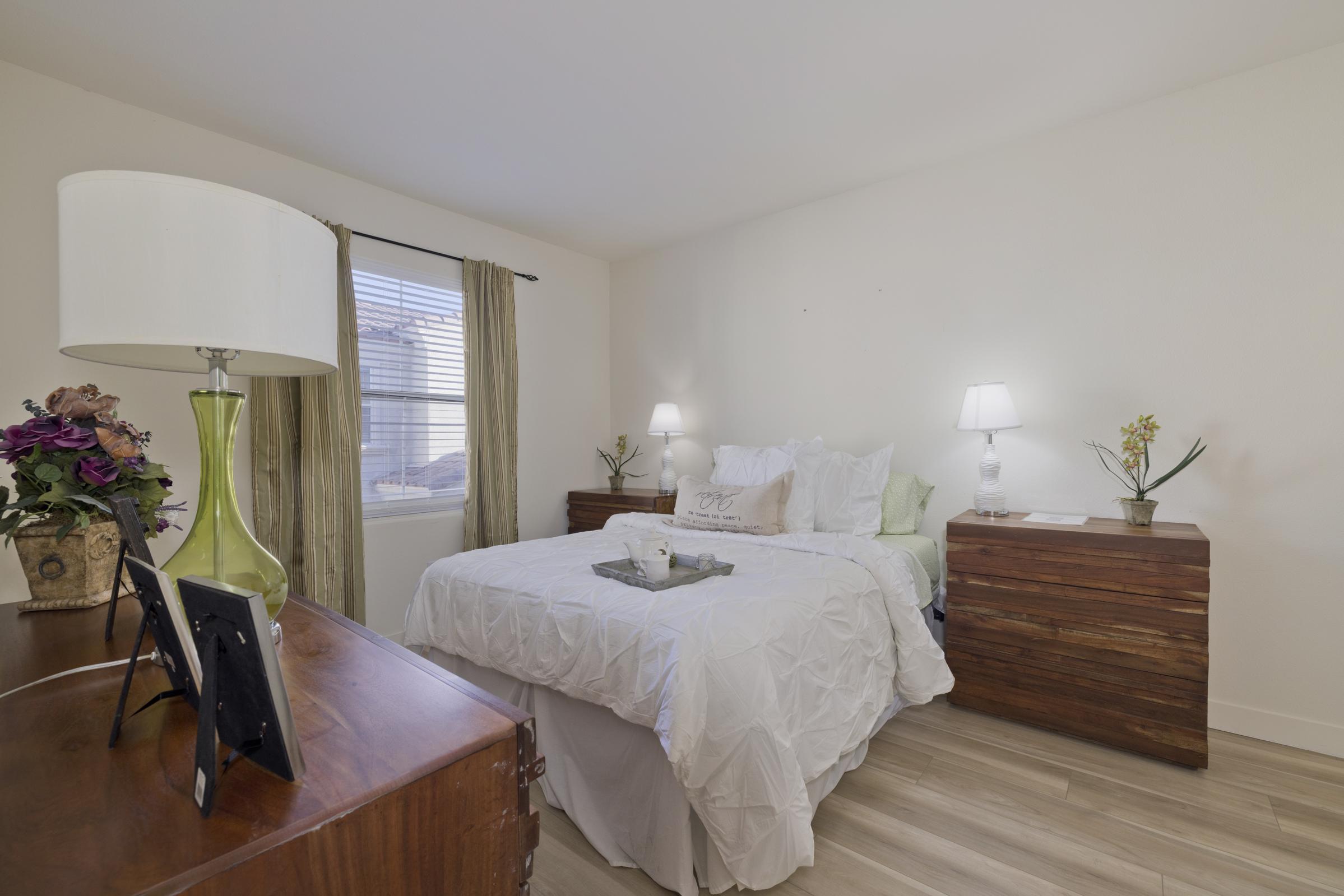 A cozy bedroom with a neatly made bed featuring white linens. Two wooden nightstands with lamps are on either side of the bed. A window with light curtains allows natural light to enter the room. A decorative plant and a small tray on the bed add a touch of elegance. The walls are painted a soft, neutral color.