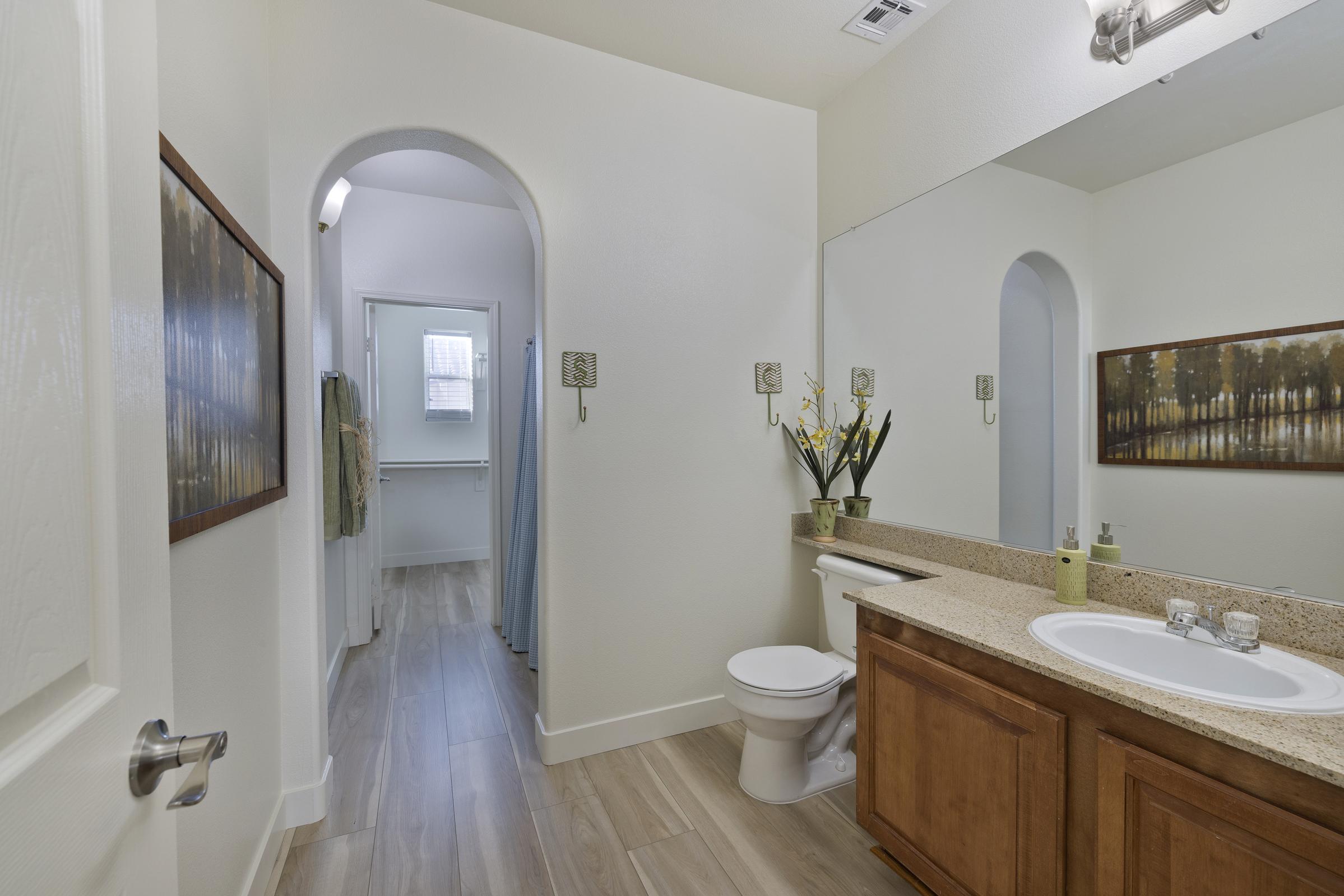 A spacious bathroom featuring a light color scheme, with a sink and a toilet. The walls are painted white, and there's a large mirror above the sink. A small round window allows natural light. Decorative elements include a wall painting and a potted plant on the counter. The floor is tiled with light wood-like flooring.