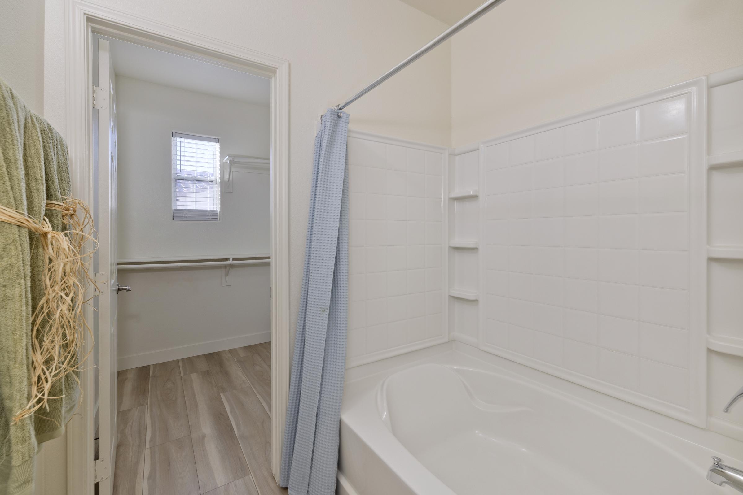 A clean bathroom featuring a white bathtub and tiled walls. A blue shower curtain hangs on a rod, and there are shelves for toiletries. A doorway leads to a room with natural light from a window. Soft green towels are neatly hung nearby, adding a touch of color to the neutral decor.