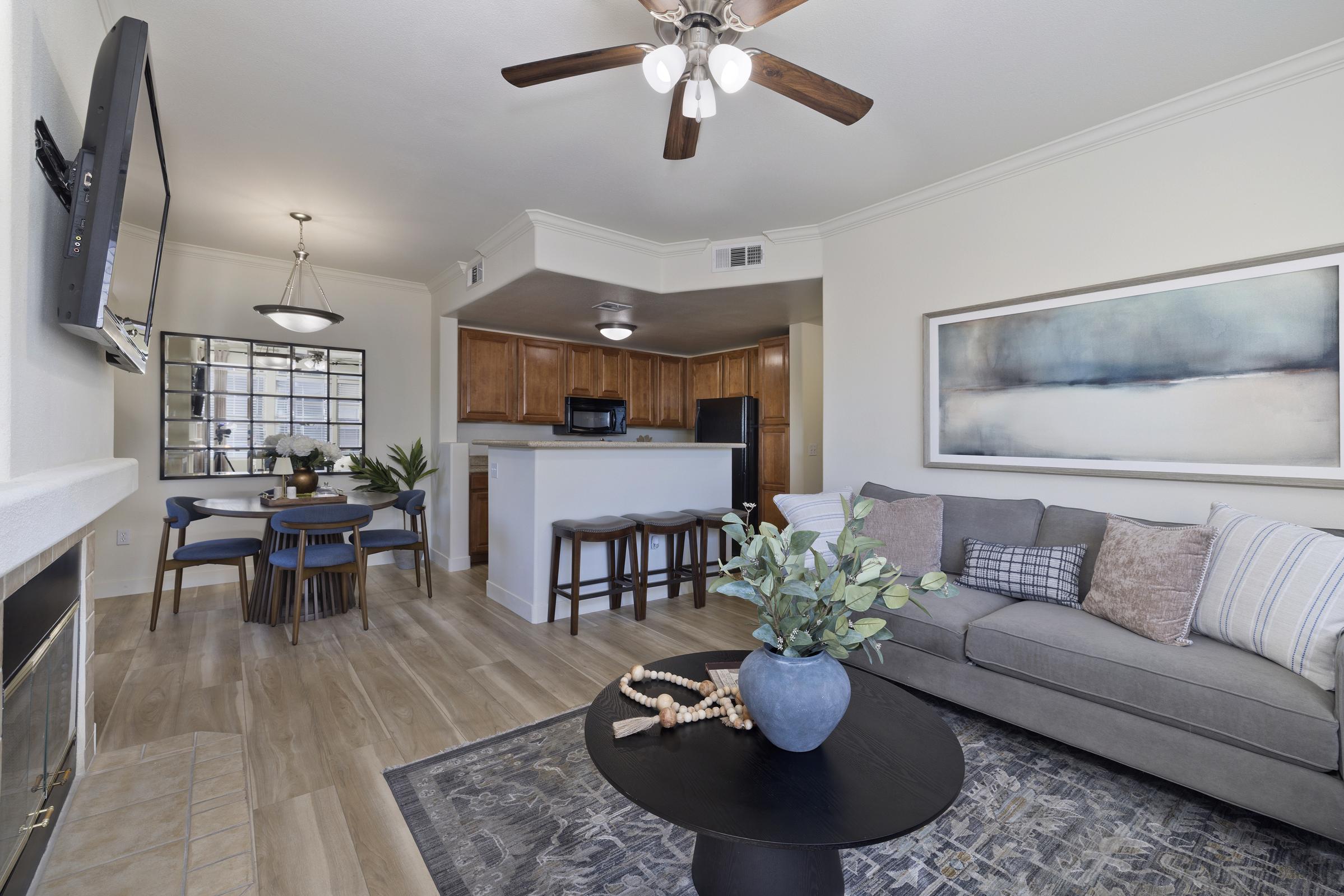 A cozy living room featuring a gray sofa with decorative pillows, a round coffee table with a plant centerpiece, and a flat-screen TV mounted on the wall. In the background, there is a dining area with a round table and four chairs, and a modern kitchen with wooden cabinets. The space is well-lit and inviting.