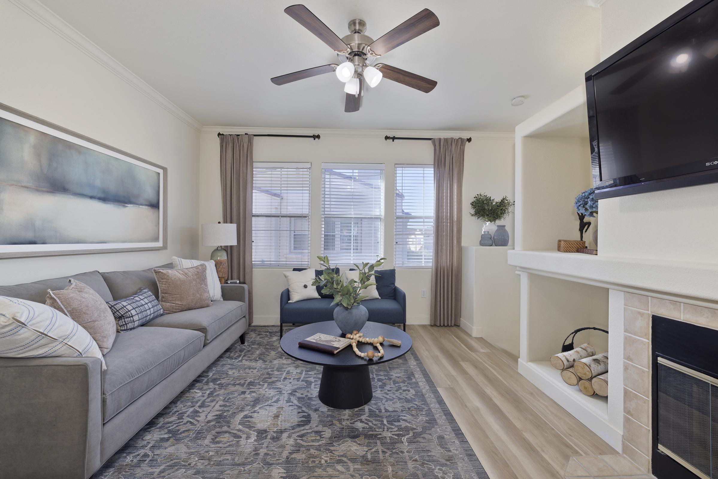 A modern living room featuring a gray sofa with decorative pillows, a round black coffee table, and a large abstract painting on the wall. Natural light streams through two windows with curtains, and a ceiling fan adds to the décor. A fireplace is visible with logs stacked next to it.