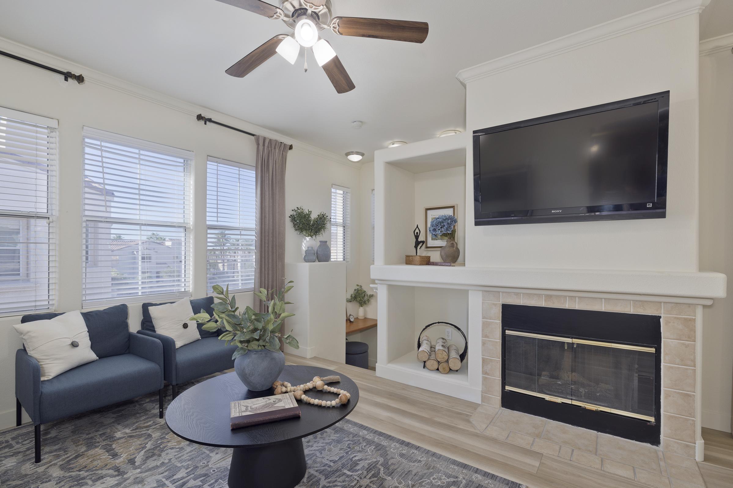 A cozy living room featuring two blue chairs, a round black coffee table with decorative items, and a fireplace. A flat-screen TV is mounted on the wall above the fireplace. Sunlight streams through several windows with white blinds, enhancing the warm ambiance of the space.