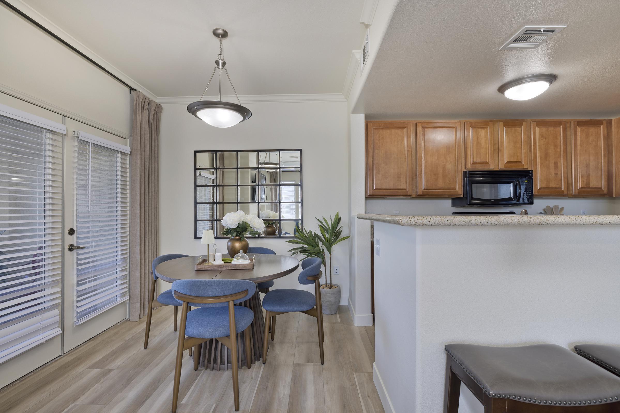 A modern dining area featuring a round wooden table with blue chairs, a centerpiece of flowers, and a mirror on the wall. Large windows with blinds provide natural light, and the kitchen area in the background has wooden cabinets and a microwave. The decor is stylish and inviting.
