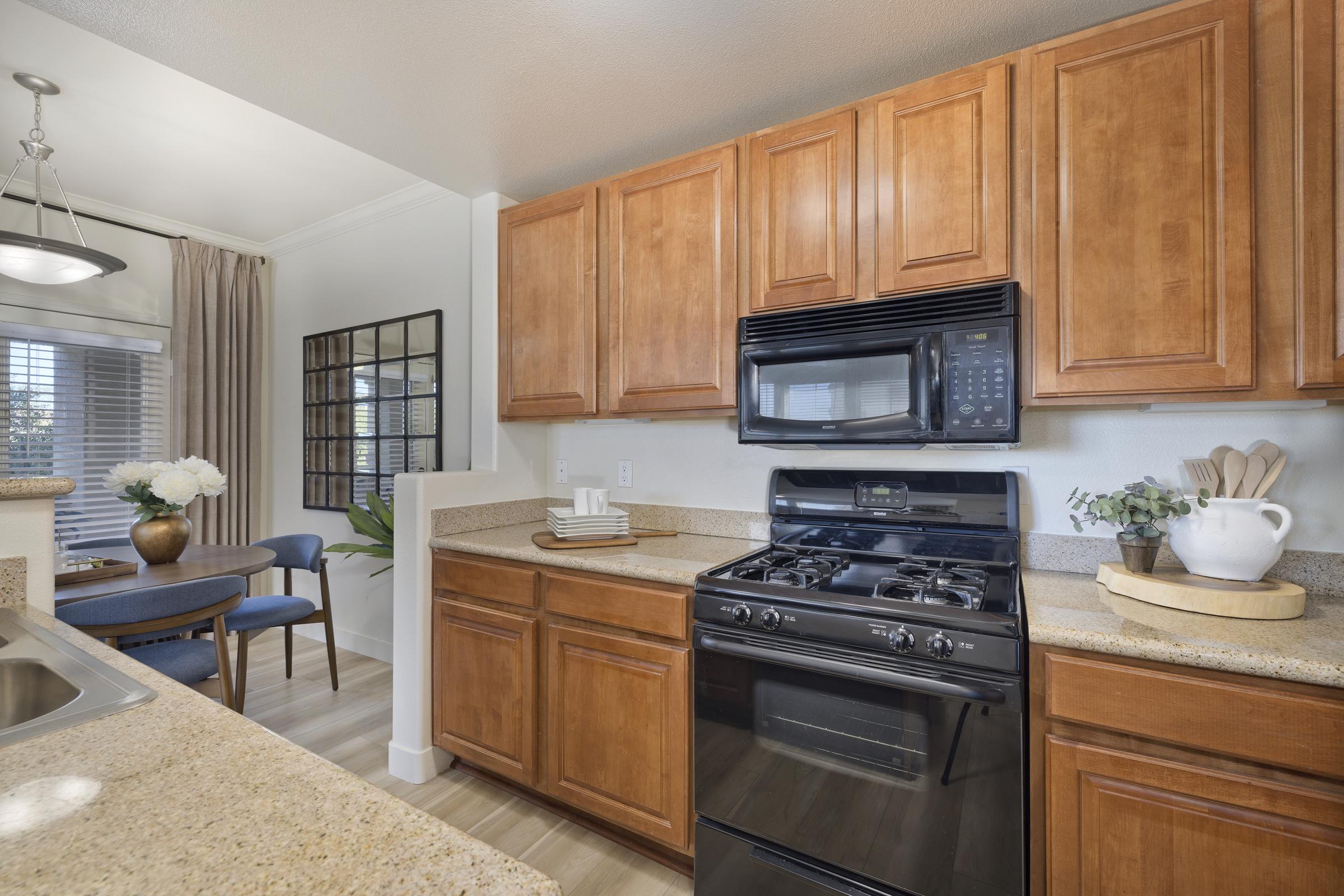 A modern kitchen featuring wooden cabinetry, a black gas stove with an oven, and a microwave above. The countertop is made of light-colored stone, with decorative items and plants. In the background, a dining area is visible with a round table and chairs, and natural light coming through a window.
