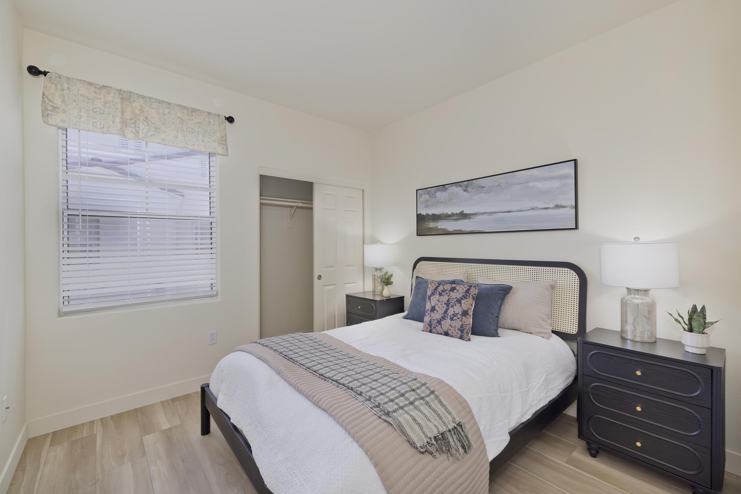 A cozy bedroom with a queen-sized bed featuring a white comforter and decorative pillows. A small nightstand with a lamp and a plant is beside the bed. There's a window with blinds and a closet door visible. The walls are painted a soft beige, and a landscape painting hangs above the bed. Wooden flooring adds warmth to the room.