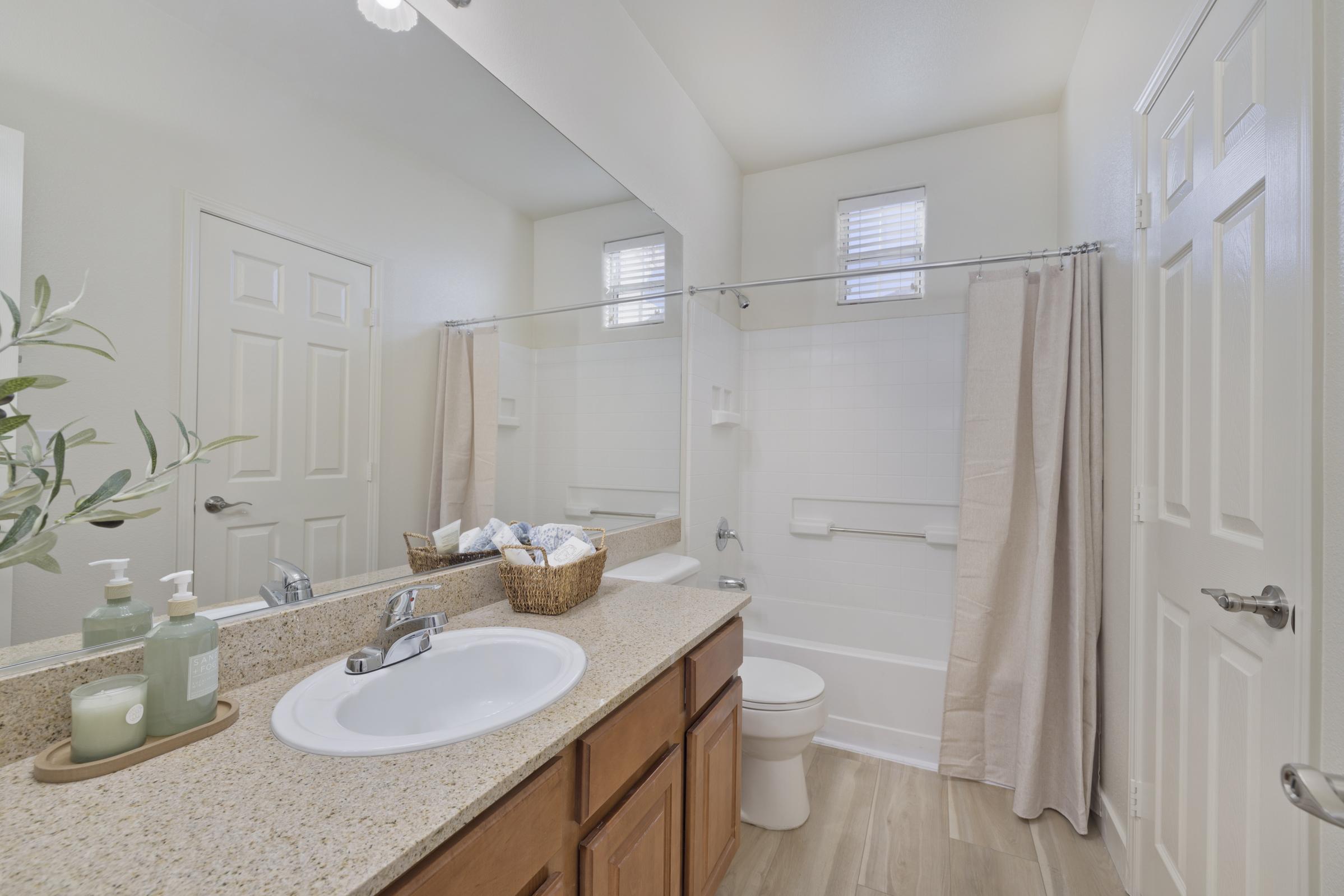 A clean and modern bathroom featuring a bathtub with a shower curtain, a large mirror, and a double sink vanity with decorative containers. The neutral-colored walls and flooring create a bright atmosphere, enhanced by natural light from small windows. Decor includes potted greenery and neatly arranged towels.