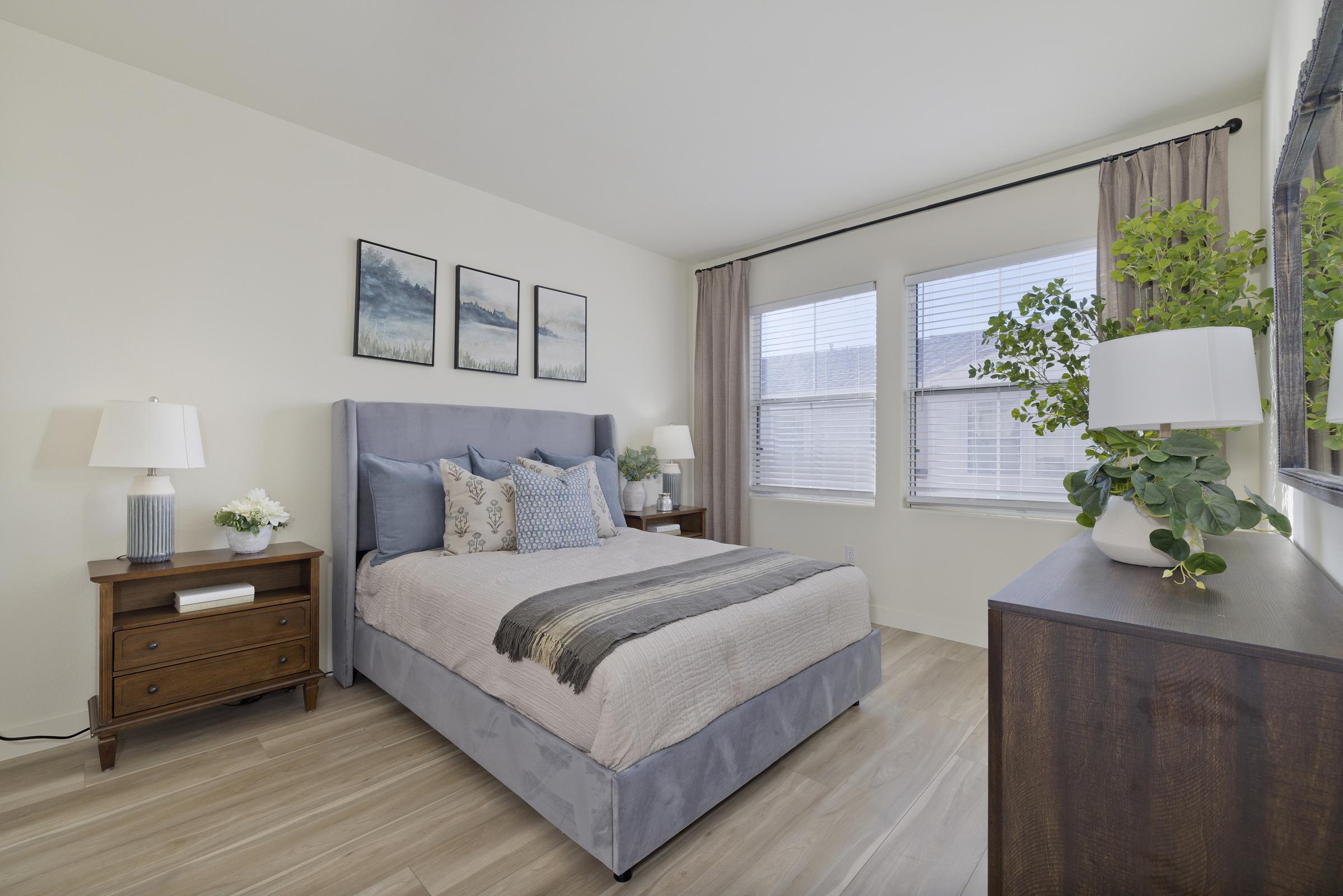 A modern bedroom featuring a neatly made bed with a soft quilt and decorative pillows. There's a wooden nightstand with a lamp and a vase of flowers beside the bed. A dresser with a plant on top is positioned against the wall, and large windows allow natural light to brighten the space. Three framed artworks adorn the wall.
