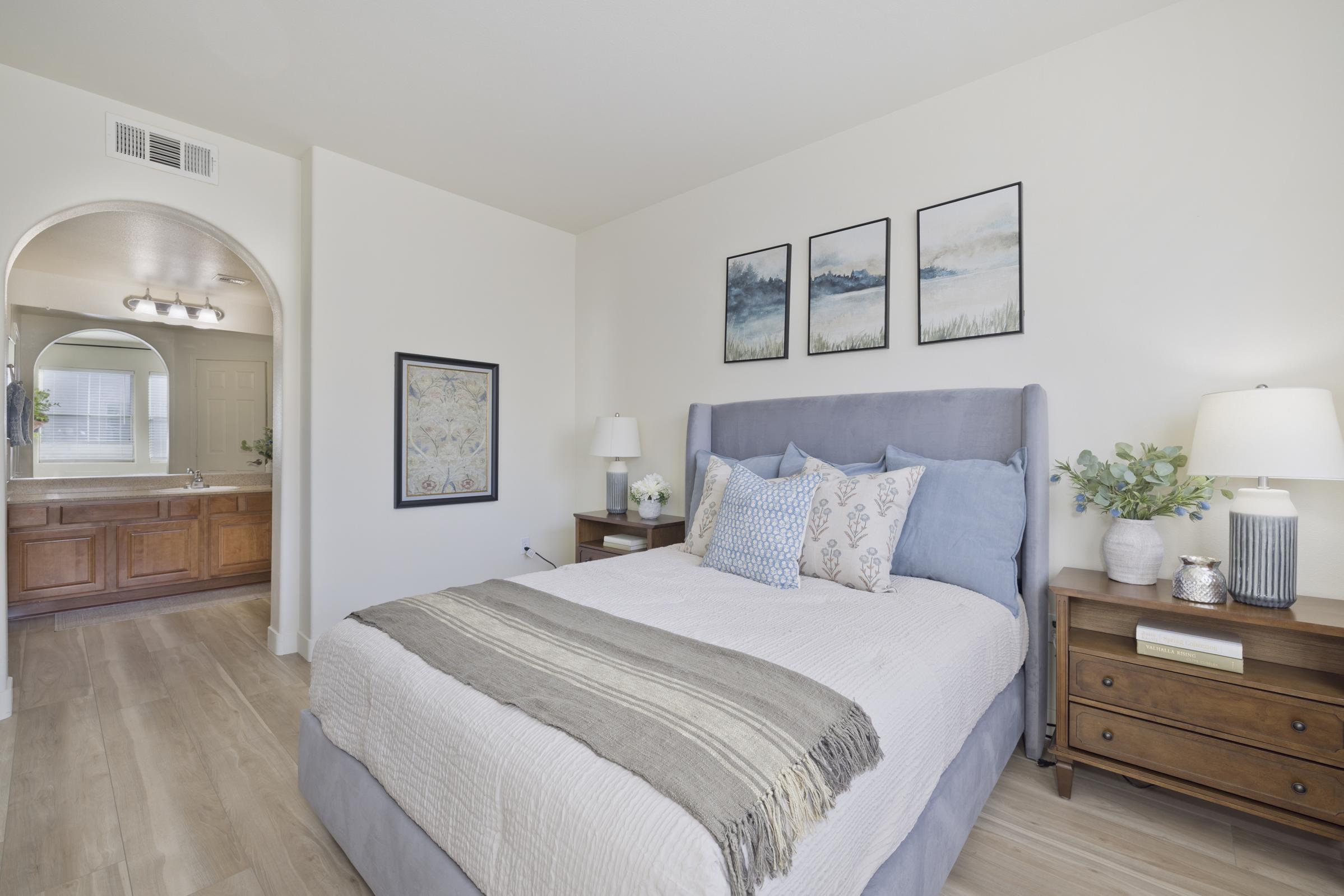 A cozy bedroom featuring a bed dressed in light-colored linens and a striped throw. Two bedside tables hold lamps and decorative plants. Three framed artworks are mounted on the wall. An arched doorway leads to a bathroom area with wooden cabinetry and a mirror. Natural light brightens the space.