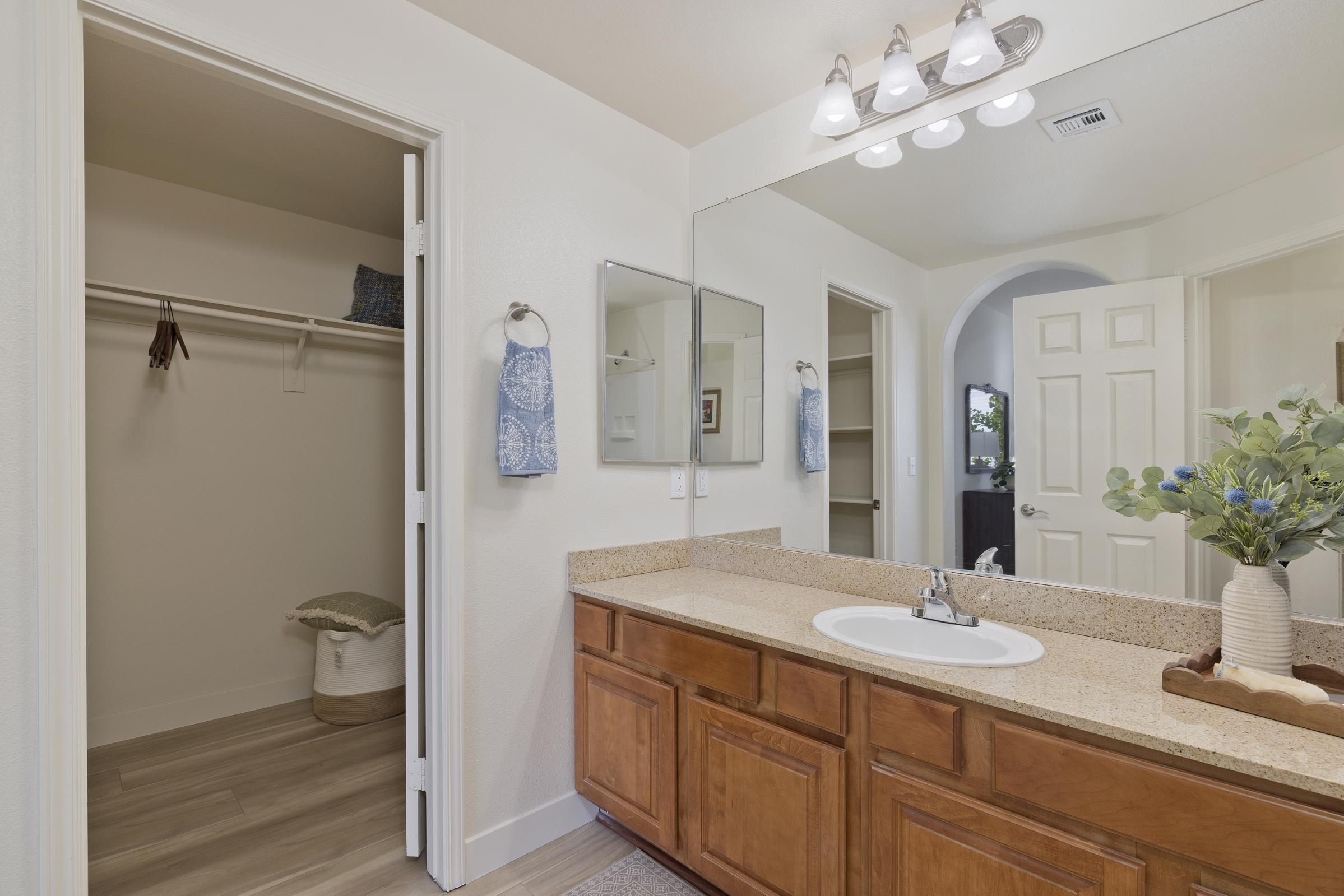 A modern bathroom featuring a double vanity with wooden cabinets and a sink, flanked by two mirrors. On the left, there's a walk-in closet with a white shelf and a storage basket. The walls are painted light, and there are decorative towels hanging by the sink. Soft lighting illuminates the space.