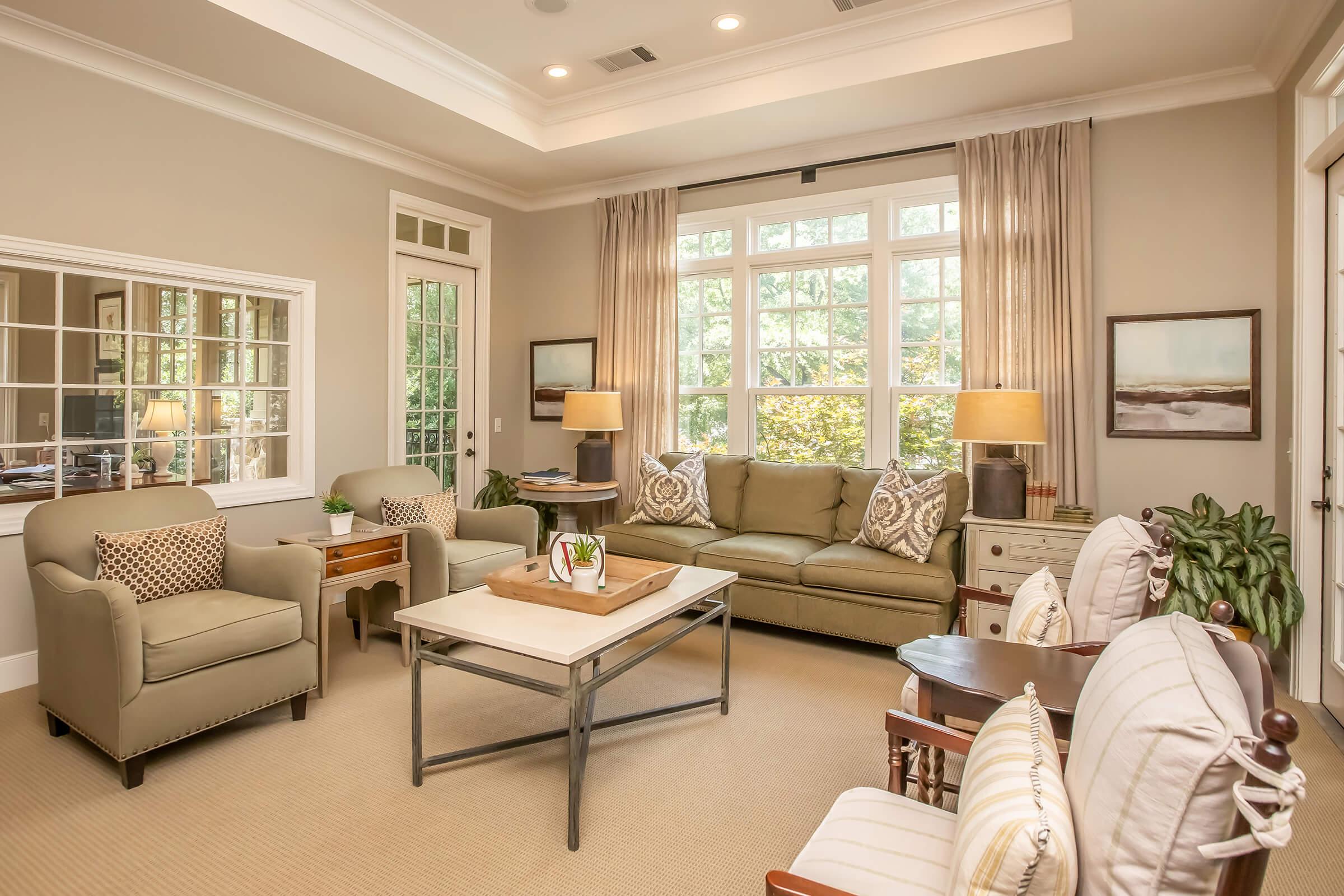 A cozy, well-lit living room featuring a light-colored sofa with decorative pillows, two armchairs, and a coffee table with decor. Large windows allow natural light to fill the space, complemented by soft curtains. A few potted plants add a touch of greenery, and a mirror reflects the room's inviting atmosphere.