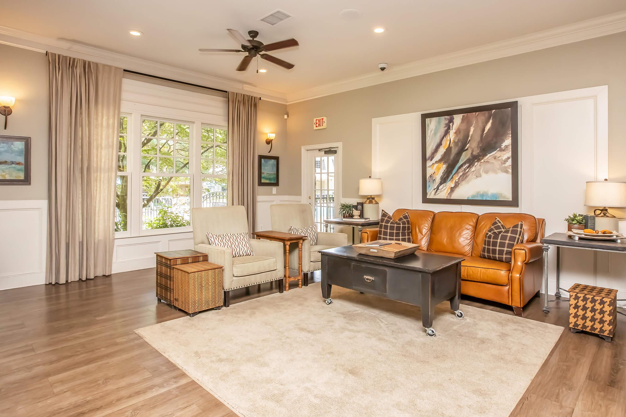 A cozy living room with hardwood floors, featuring a brown leather sofa, two light-colored armchairs, and a large area rug. There’s a black coffee table in the center, a side table with a lamp, and large windows letting in natural light. Decorative paintings and plants add warmth to the space.