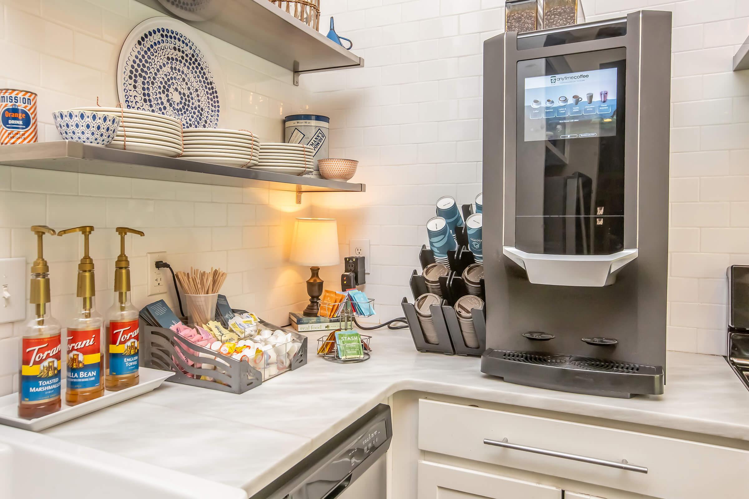 A modern kitchen countertop featuring a beverage dispenser with touch screen controls, surrounded by coffee supplies including flavored syrups and cups. Shelves above display assorted plates and kitchenware, creating a tidy and inviting atmosphere.
