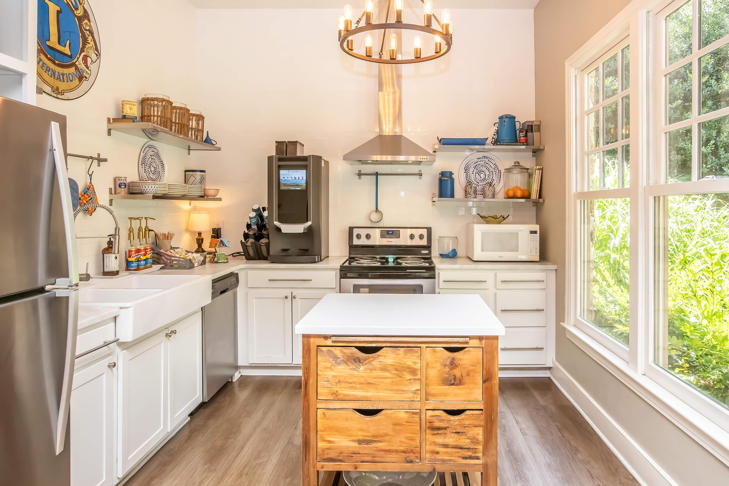 Modern kitchen featuring white cabinetry, a central wooden island with storage, stainless steel appliances including a fridge and oven, and open shelving displaying dishware. A large window offers natural light and a view of greenery outside. The space combines functionality with a cozy aesthetic.