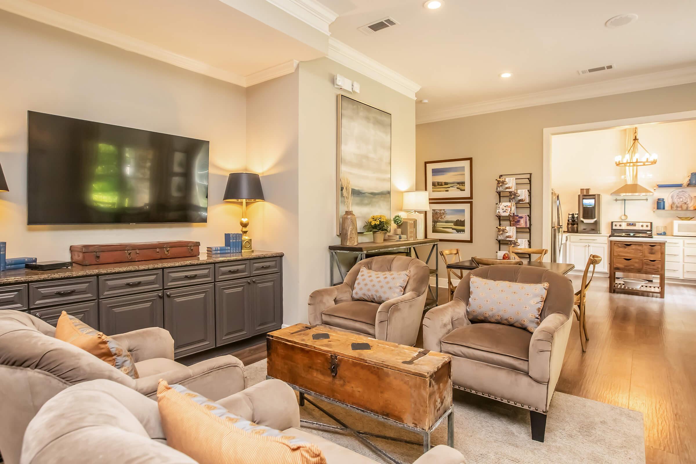 A cozy living room featuring a flat-screen TV mounted on the wall, a stylish console cabinet, and a vintage coffee table. Two comfortable armchairs with cushions are arranged around the table, with a warm color palette and soft lighting creating an inviting atmosphere. A doorway leads to a bright kitchen in the background.