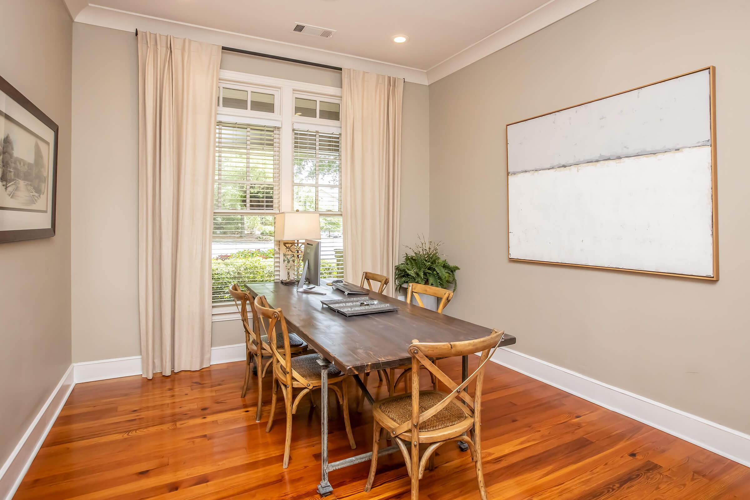 A well-lit dining room featuring a wooden table surrounded by four chairs, a lamp on the table, a plant in the corner, and large windows with sheer curtains. The walls are painted a soft beige, and there's a minimalist artwork on one wall, creating a cozy and inviting atmosphere.