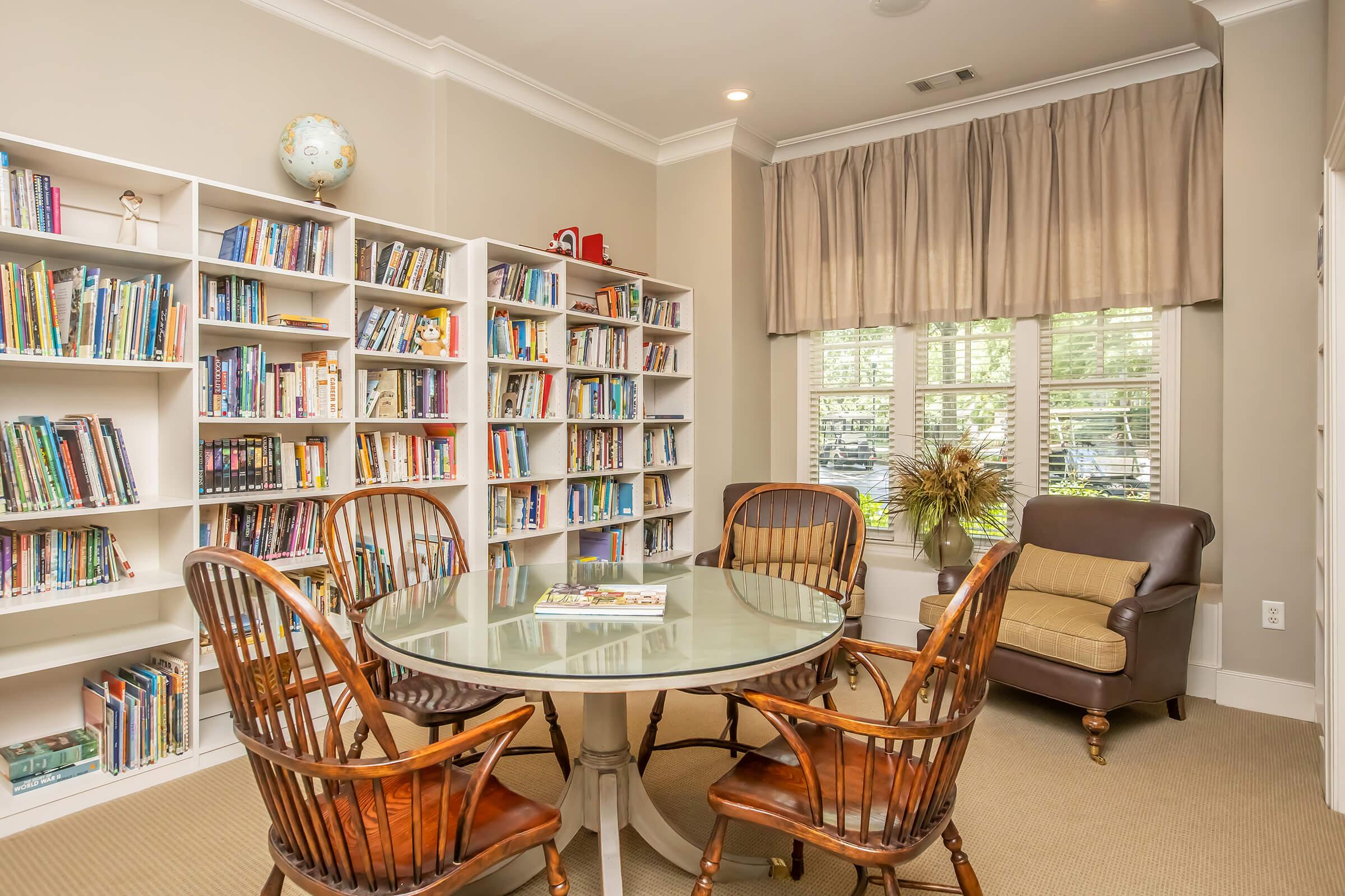 A cozy reading room featuring a round glass table surrounded by four wooden chairs. Shelves filled with books line the walls, and a comfortable armchair is positioned near a window with soft natural light filtering in through sheer curtains. A globe sits atop a shelf, adding a decorative touch.