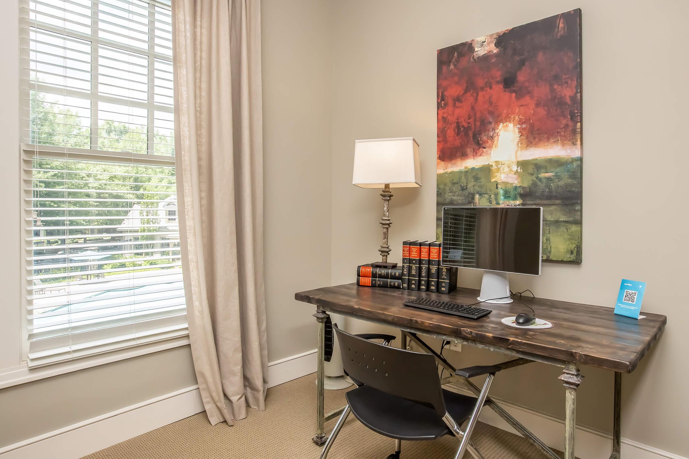 A modern workspace featuring a wooden desk with a computer monitor, keyboard, and accessories. Behind the desk, there is a lamp and a large abstract painting on the wall. The room has natural light coming through a window with blinds and is decorated in neutral tones.
