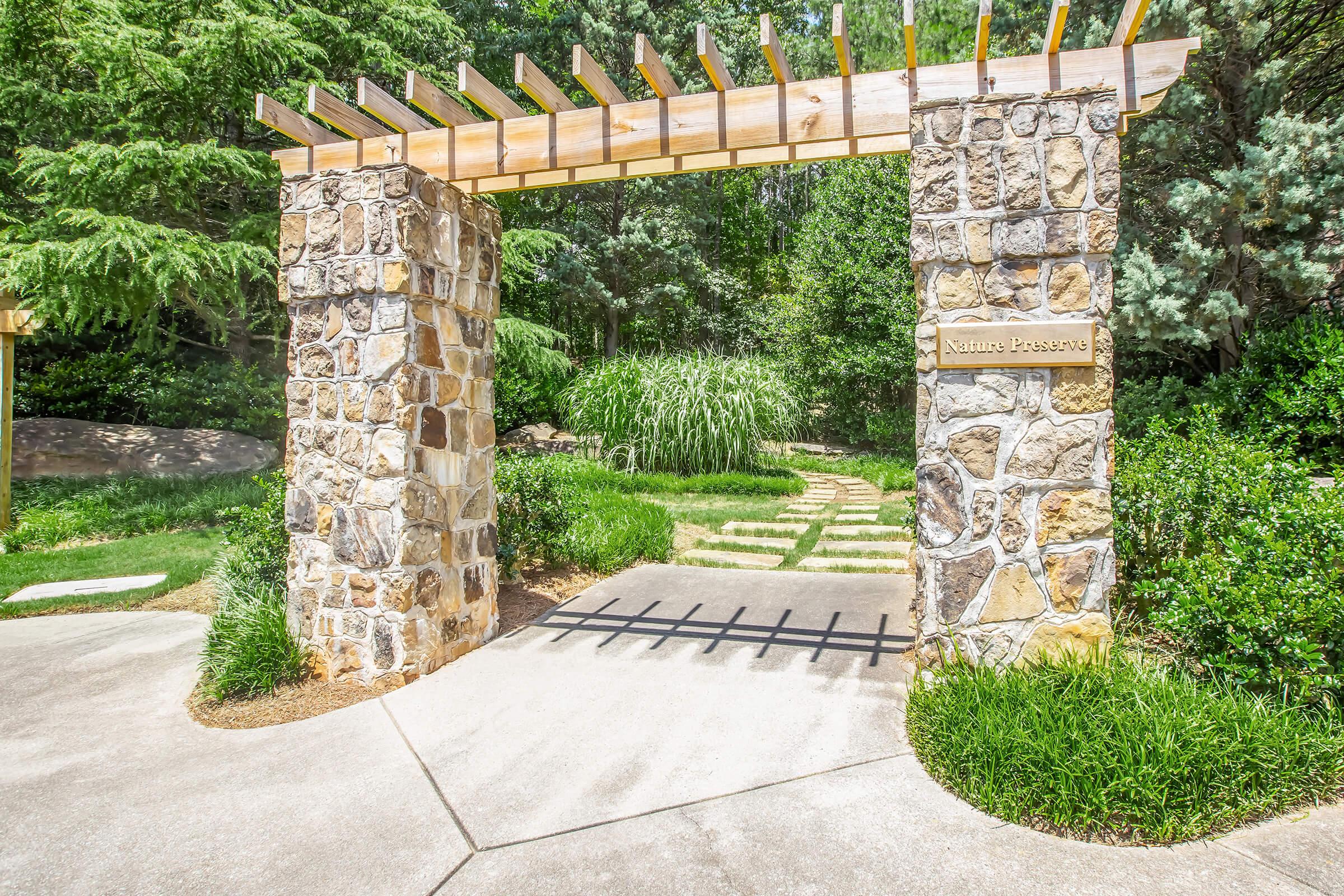 A stone archway with wooden beams leads into a lush garden. The entrance is surrounded by greenery, including tall grasses and shrubs. A pathway can be seen beyond the entrance, inviting exploration of the serene natural environment.