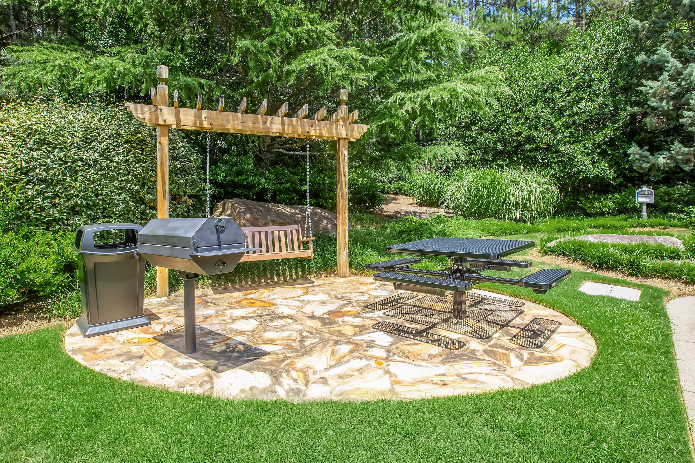 An outdoor picnic area featuring a charcoal grill, a wooden swing set, and a black picnic table on a stone patio surrounded by lush greenery and trees. The setting offers a cozy space for gatherings or relaxation in a natural environment.