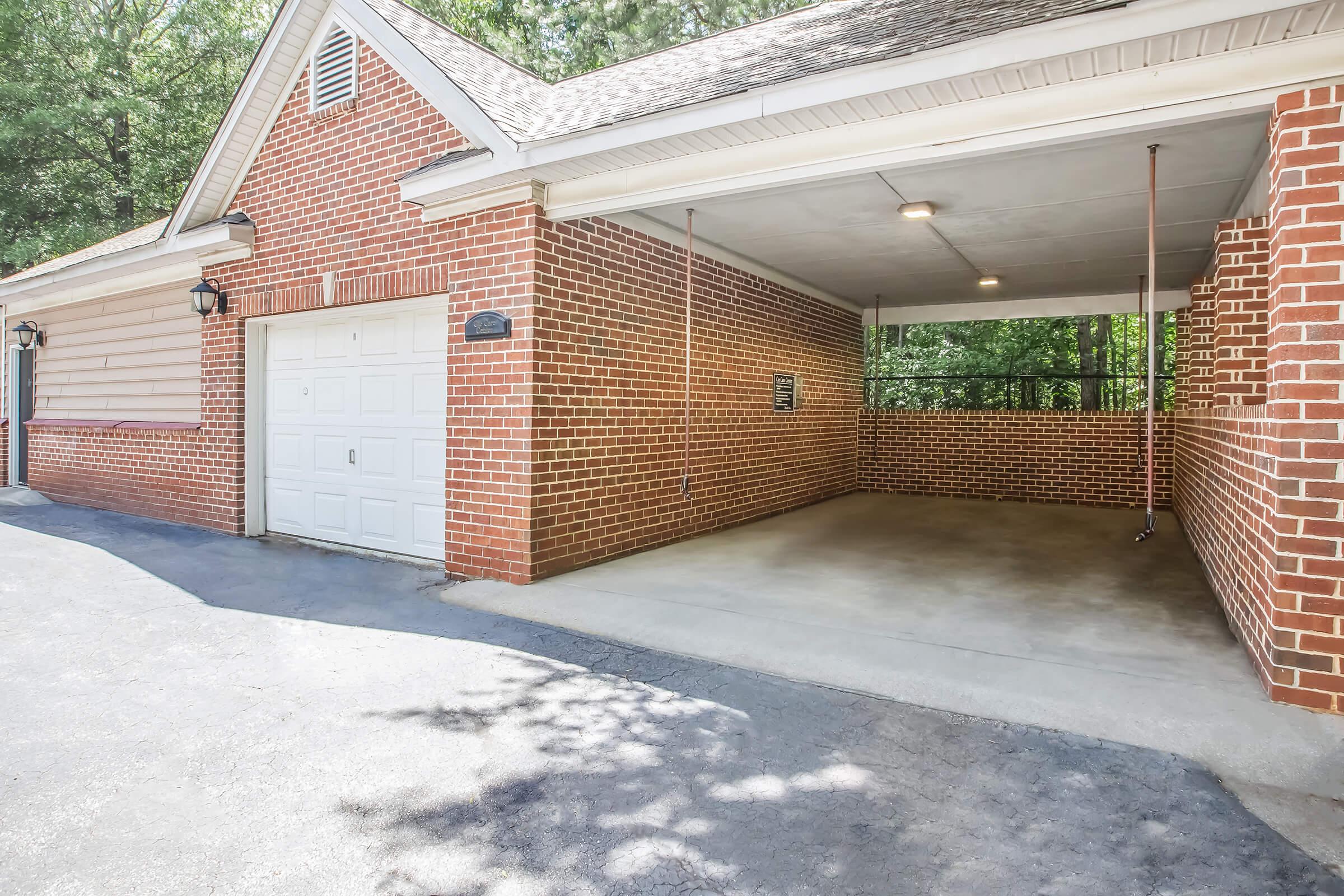 A garage with a brick exterior featuring a white garage door, empty interior space, and overhead lighting. The area is clean and well-maintained, surrounded by green trees visible through the open entrance.
