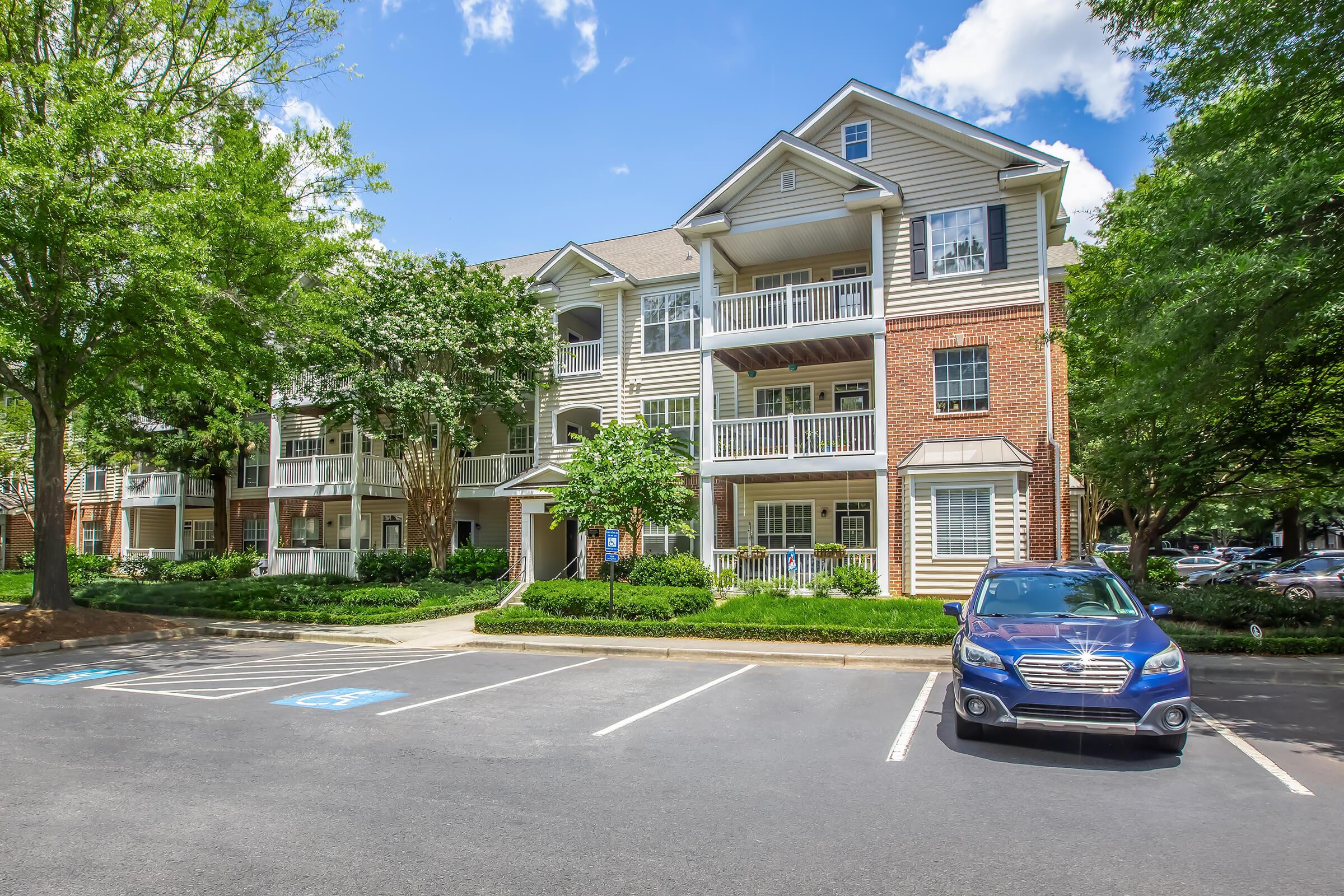 A modern multi-story apartment building with a mix of brick and siding exteriors, surrounded by lush trees and greenery. Several parked cars in the foreground, with one blue vehicle prominently displayed in a designated parking space. Bright blue sky with scattered clouds provides a sunny atmosphere.