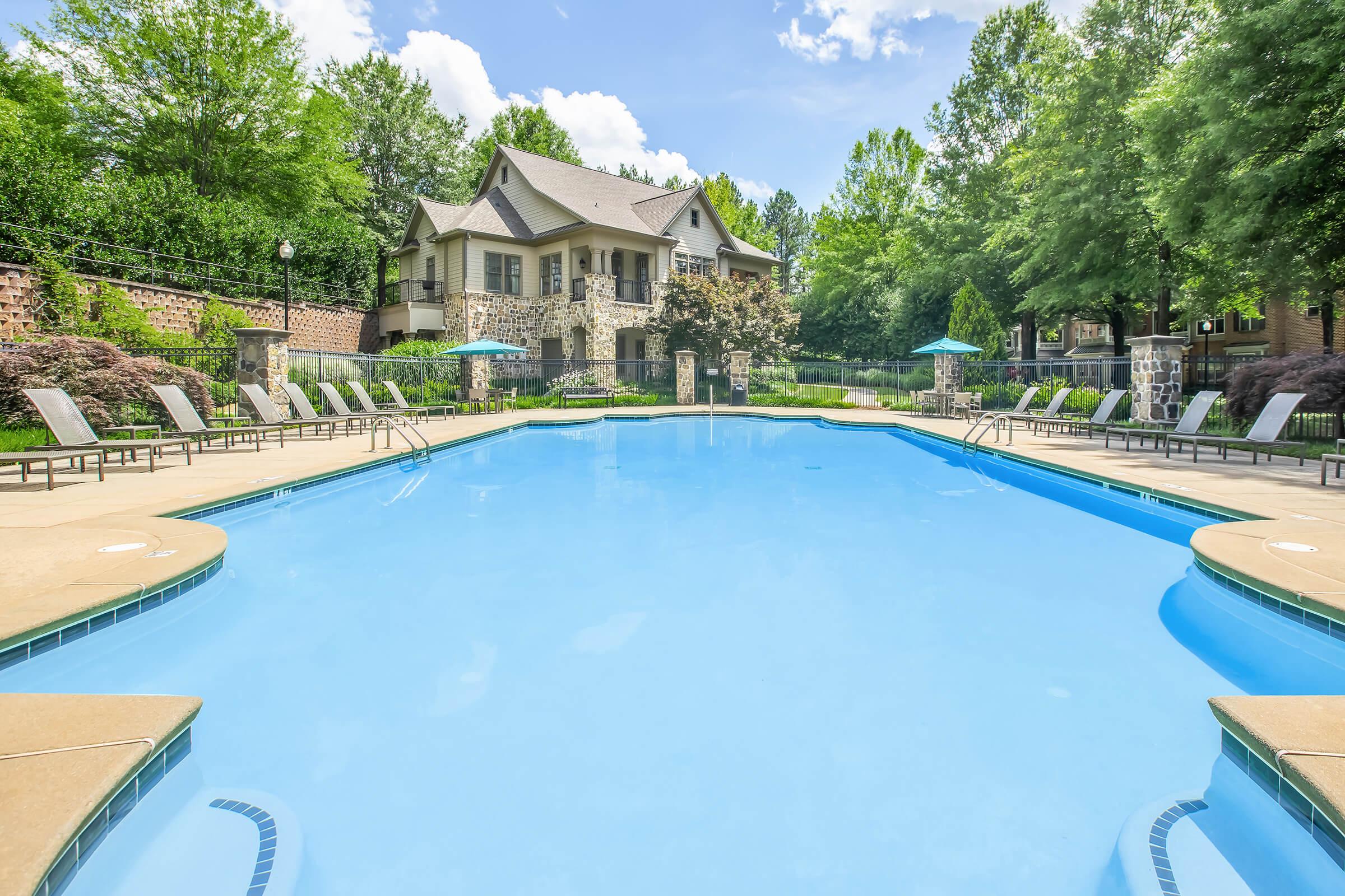A clear blue swimming pool surrounded by lounge chairs and shaded by trees, with a large stone house in the background. The sky is bright and partly cloudy, creating a serene and inviting outdoor space perfect for relaxation.