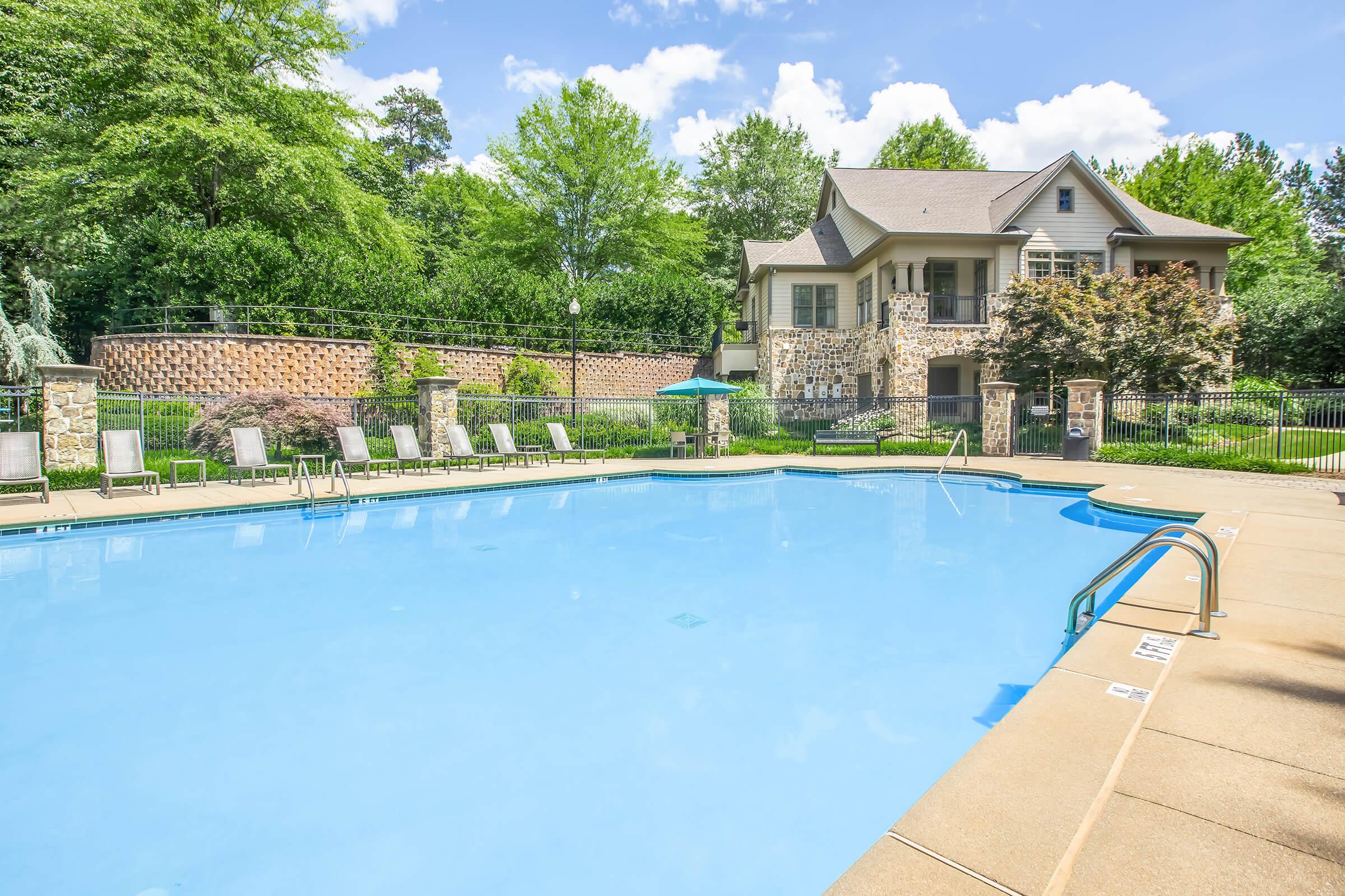 A clear blue swimming pool surrounded by lounge chairs, with a well-maintained courtyard and a stone building in the background. Lush green trees and shrubs provide a natural setting under a bright blue sky with scattered clouds.