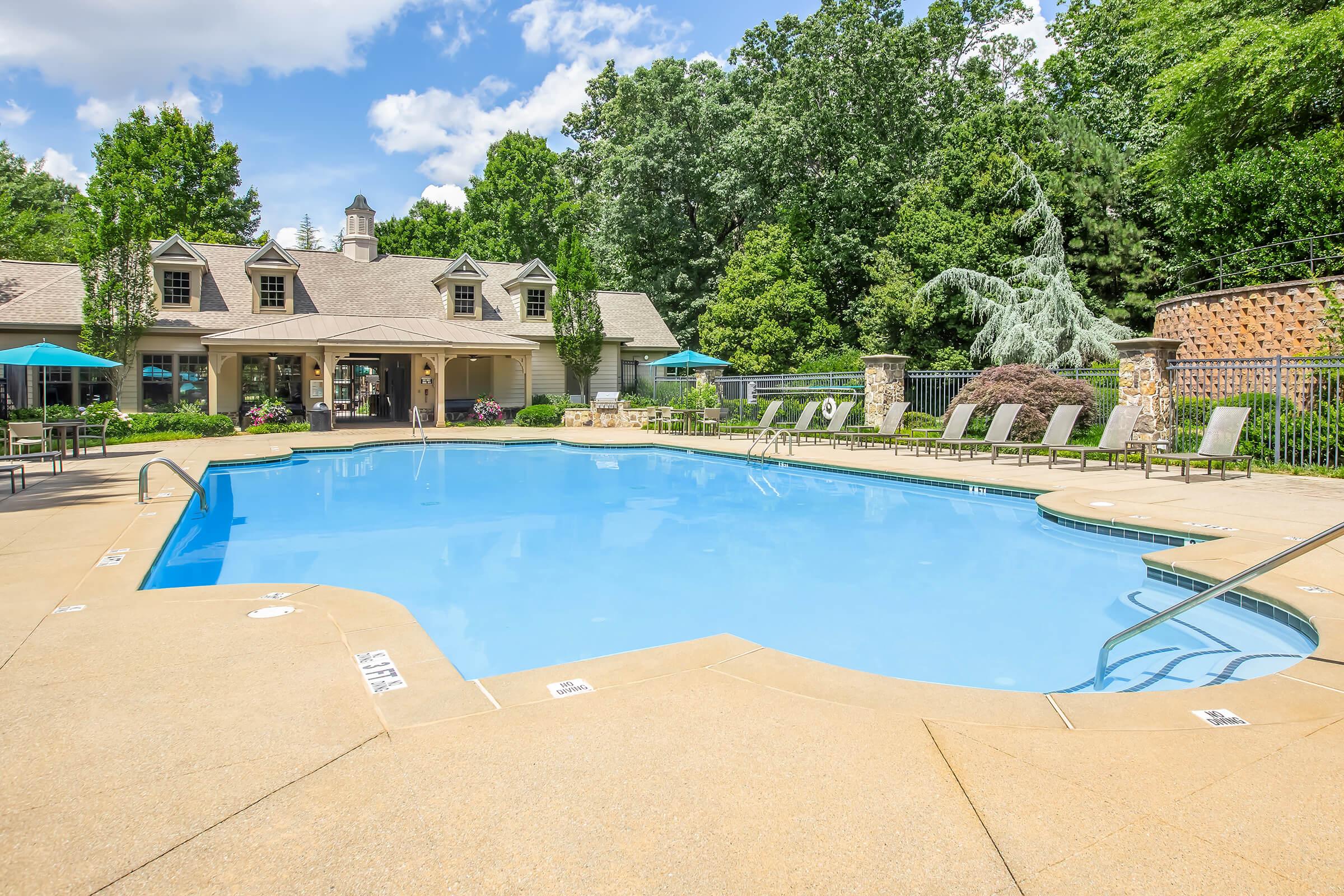 A spacious outdoor swimming pool with a clear blue surface surrounded by lounge chairs. In the background, there is a welcoming clubhouse with large windows and a shaded area featuring umbrellas. Lush green trees and landscaping frame the pool area, creating a serene and inviting atmosphere.