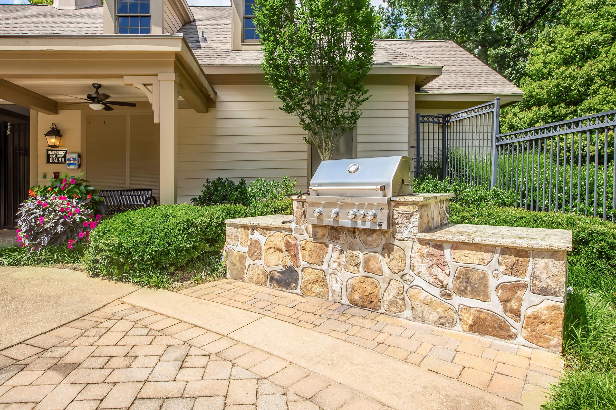 A meticulously designed outdoor grilling area featuring a stainless steel grill set on a stone countertop. The space is surrounded by greenery and colorful flowers, with a well-maintained pathway leading up to a charming house. A black iron fence adds to the cozy ambiance of the backyard.