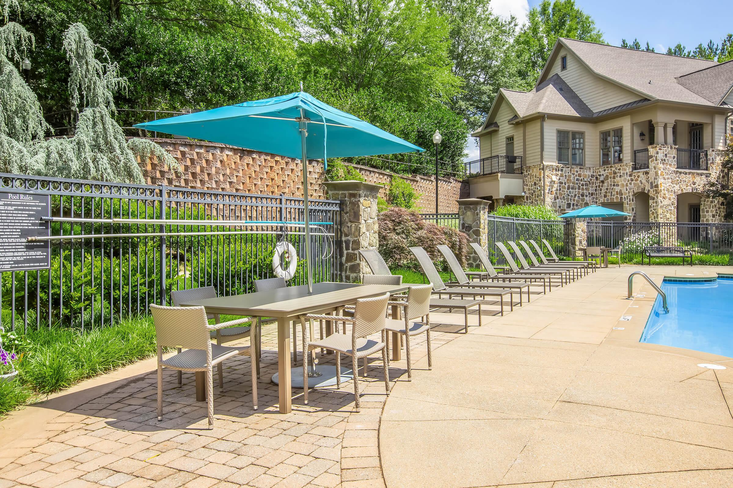 A serene poolside area featuring a sparkling blue swimming pool, surrounded by lounge chairs and a dining table with umbrellas. Lush green trees and a stone wall create a peaceful backdrop, enhancing the relaxing atmosphere for outdoor leisure.