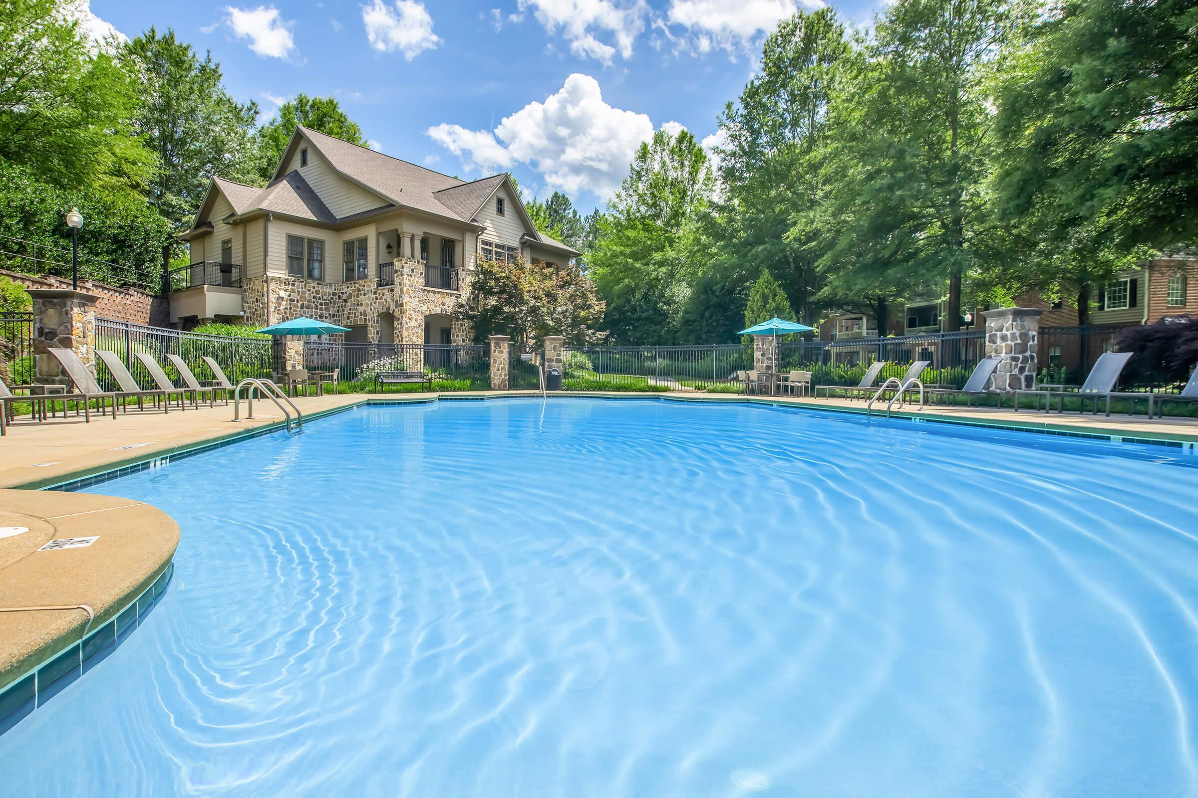 A serene outdoor swimming pool surrounded by lounge chairs, with a backdrop of a well-maintained building and lush green trees under a partly cloudy blue sky. The water is calm and inviting, reflecting the vibrant natural surroundings.