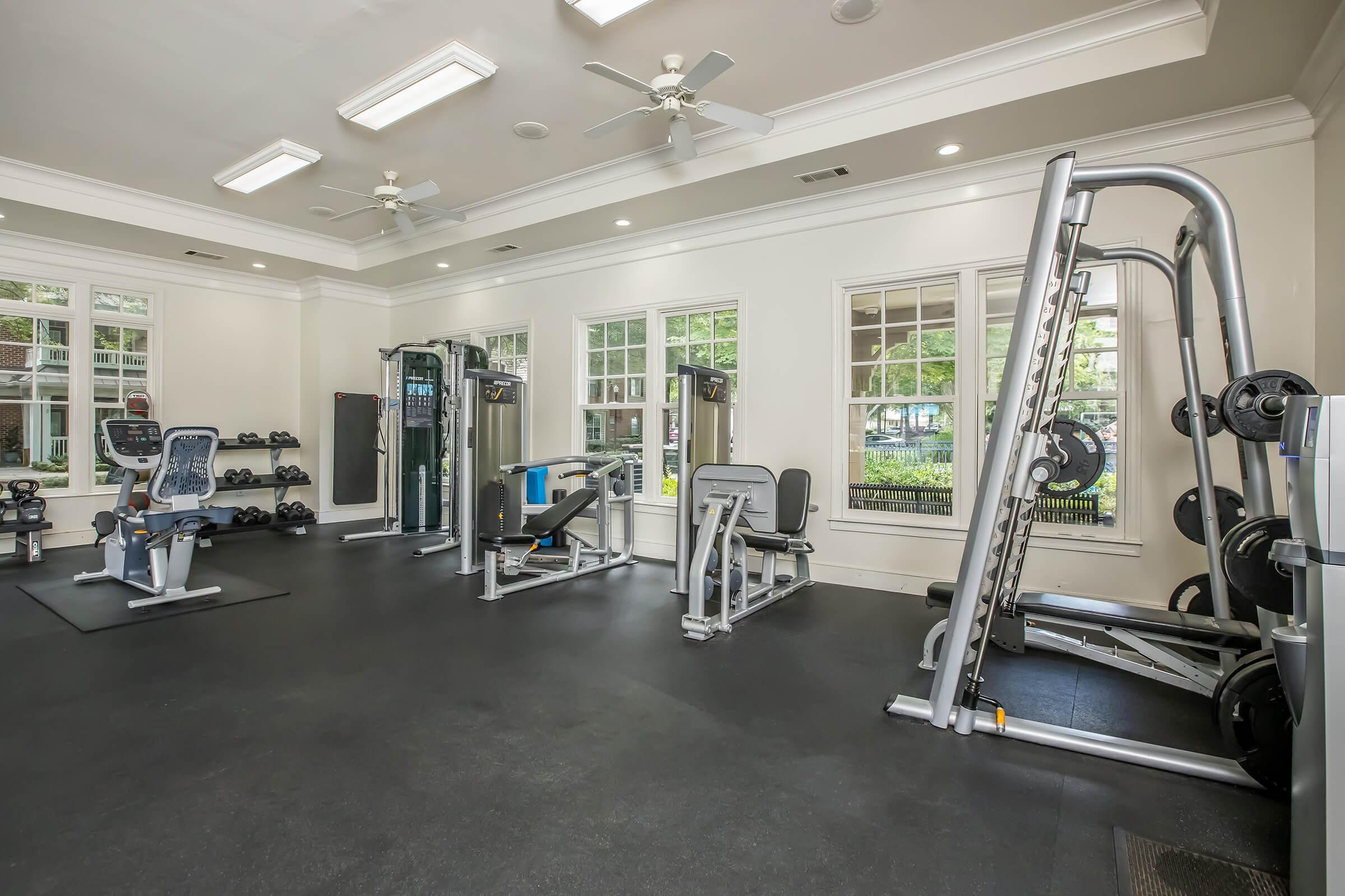 A well-equipped gym featuring various exercise machines and free weights. The space is bright with large windows allowing natural light, and includes a treadmill, weight lifting equipment, and an organized rack of dumbbells on one side. The flooring is black rubber, providing a suitable workout environment.