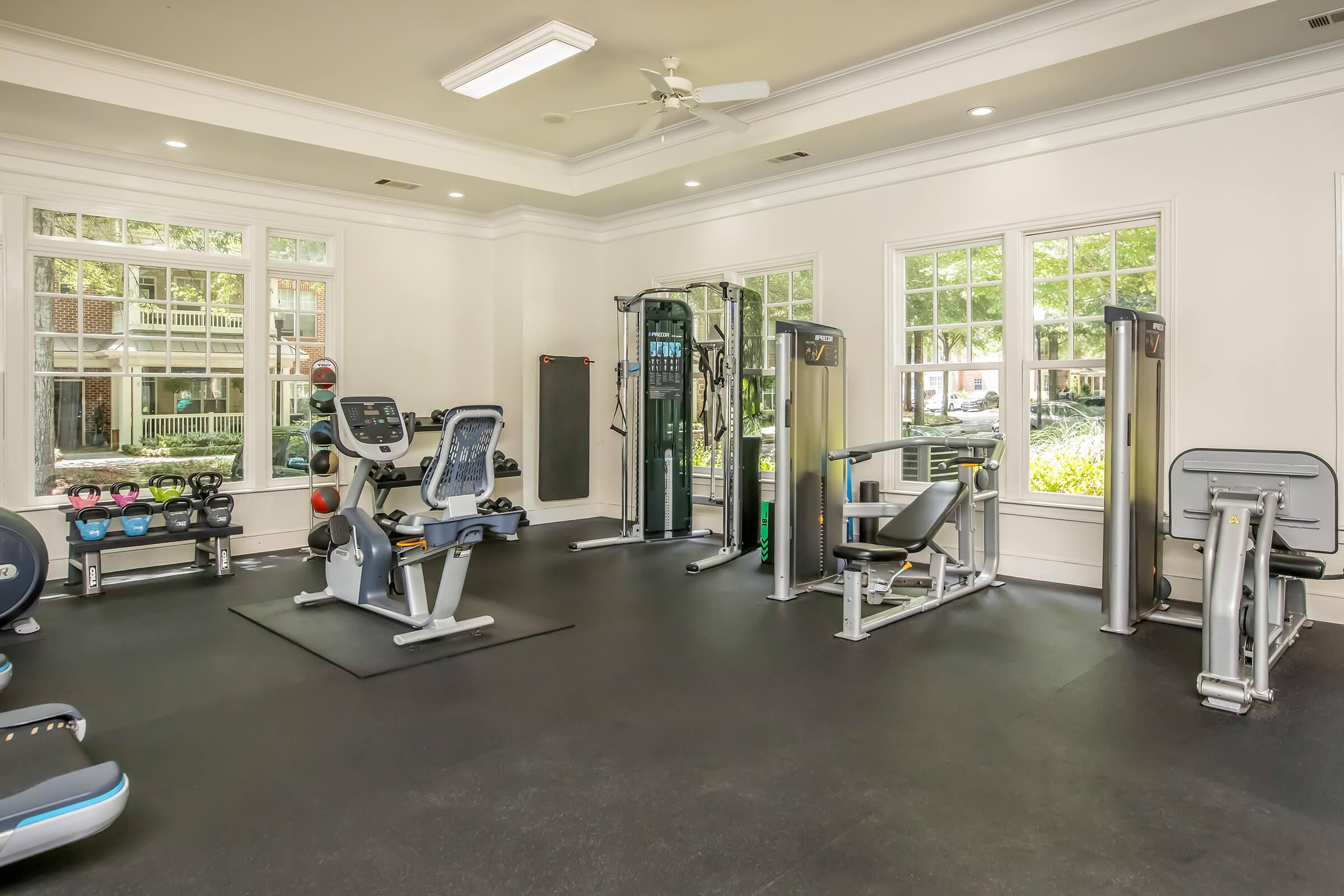A bright, spacious gym with multiple workout machines, including weightlifting equipment and cardio machines. Large windows allow natural light to fill the room, enhancing the inviting atmosphere. The floor is a dark, rubber surface suitable for exercise.