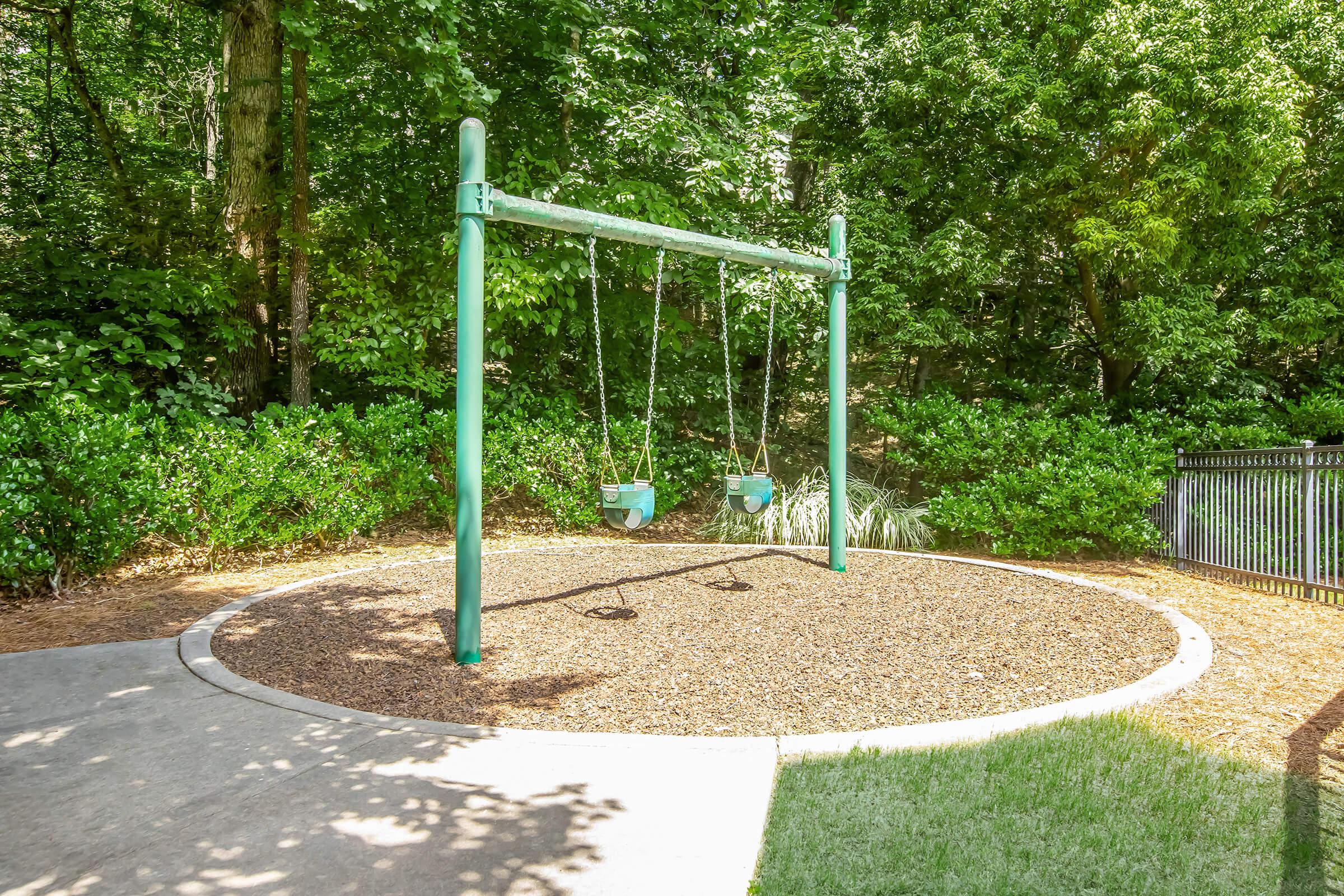 A playground area featuring a green swing set with two swings, surrounded by a mulch surface and lush green shrubbery. The setting is well-lit by natural sunlight, with a glimpse of a grassy area nearby and a fence in the background. Ideal for outdoor play and recreation.