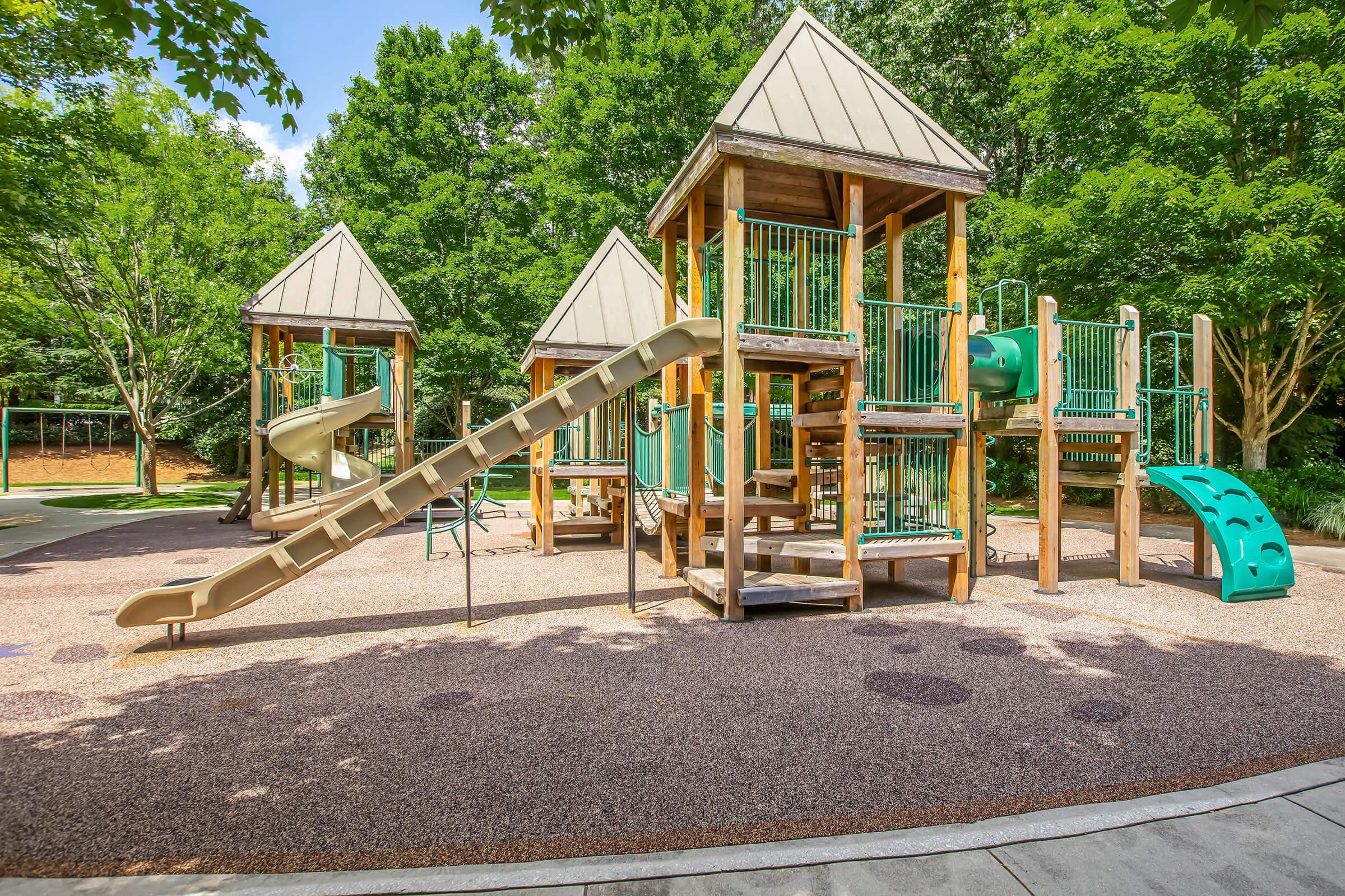 A children's playground featuring multiple play structures with slides, climbing areas, and shaded by green trees. The surface is covered with a soft, sandy material, creating a safe play environment for kids. Brightly colored equipment encourages imaginative play and outdoor activities.