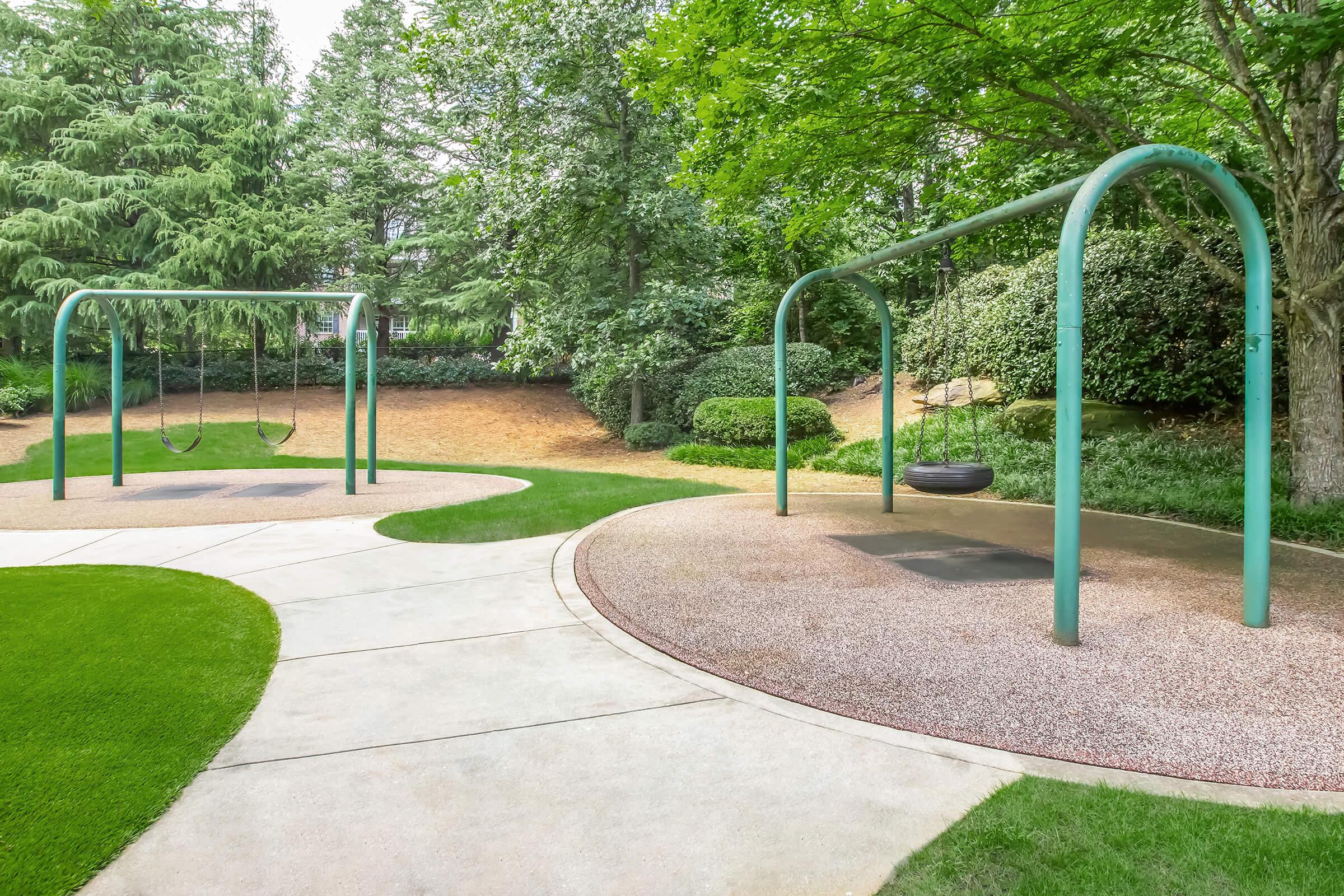 Two swings are positioned in a serene playground setting, surrounded by lush greenery and trees. The ground is covered with a smooth, pebbled surface, and a winding pathway leads through the area, creating a peaceful atmosphere for children to play and enjoy the outdoors.