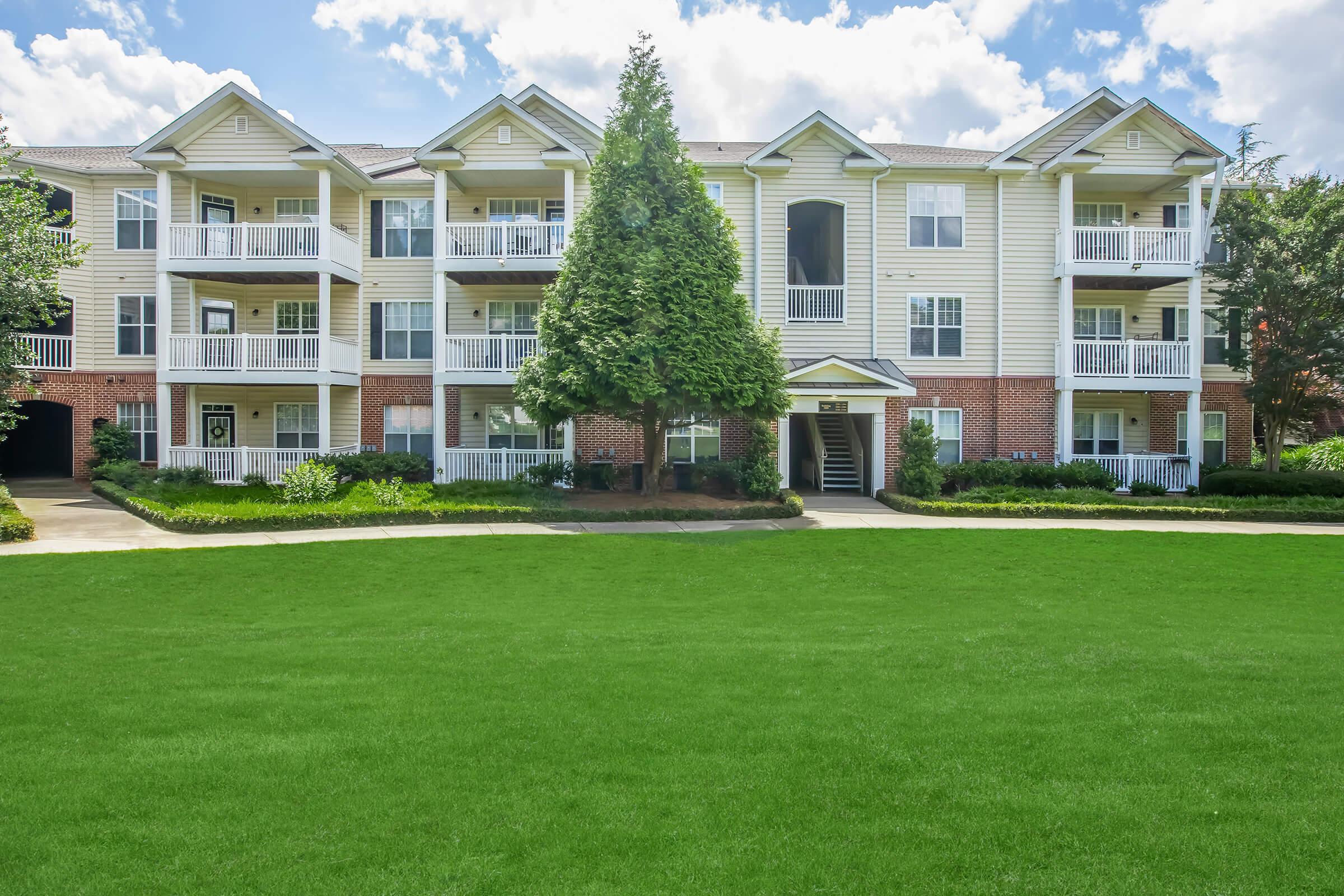 A modern apartment building with multiple balconies, surrounded by vibrant green grass and a well-maintained landscape. The exterior features a mix of brick and light-colored siding, with a central entrance and a large tree in front, under a clear blue sky with scattered clouds.