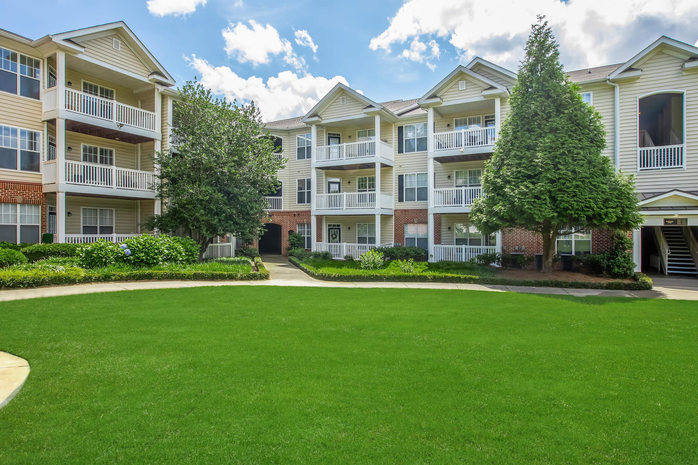 A sunny view of an apartment complex featuring three-story buildings with balconies and white railings. The well-maintained grounds include lush green grass and shrubs, with a pathway leading through the center. Bright blue sky with fluffy clouds completes the scene.