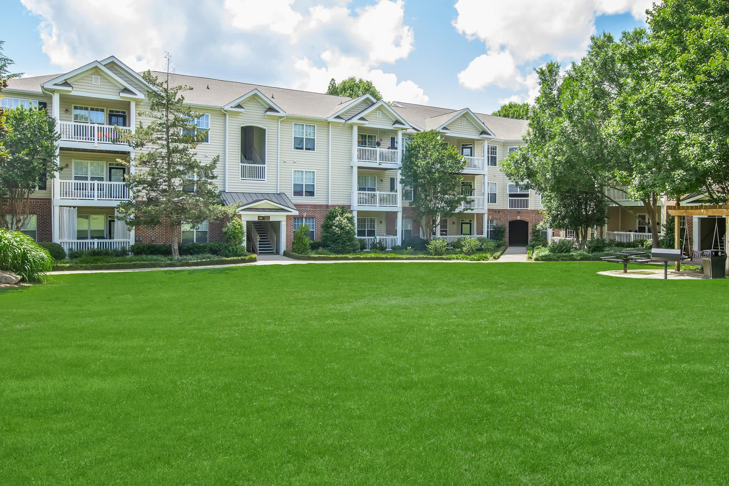 A well-maintained apartment complex featuring multiple buildings with balconies, surrounded by lush greenery and a large, manicured lawn. The sky is bright with fluffy clouds, adding to the welcoming atmosphere of the residential area.